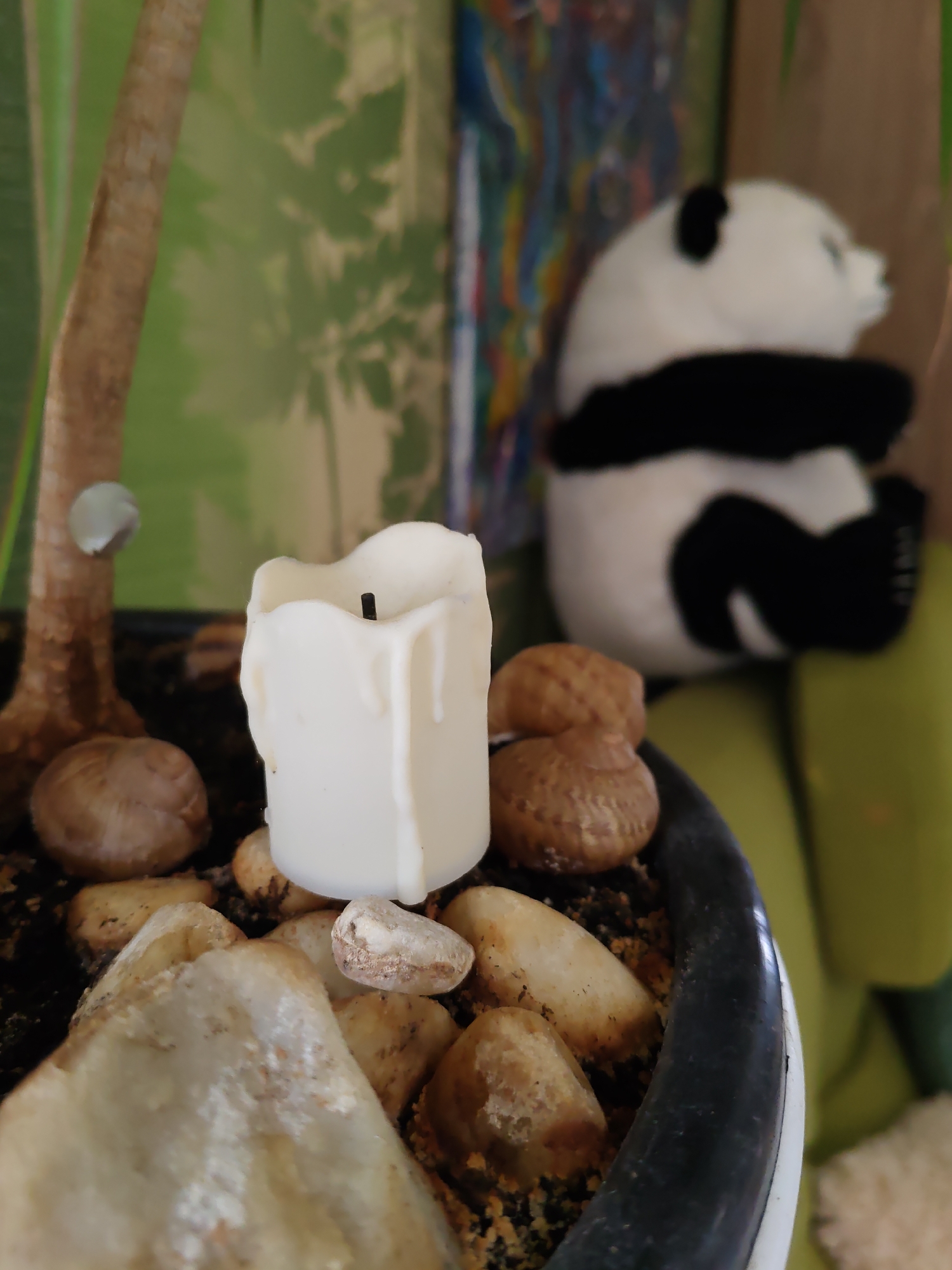 A close-up, eye-level, medium shot captures a white, dripped candle standing upright in a black planter filled with soil and assorted pebbles. To the left of the planter, a portion of a brown, textured tree trunk is visible. In the background, a blurry stuffed panda sits on a bright green surface. The wall behind the panda has a green, patterned wallpaper with shadowy, leaf-like shapes, and to the right, a section of a colorful, abstract painting can be seen. The image appears to be taken indoors, likely in a residential setting within Rostov-on-Don, Russia.