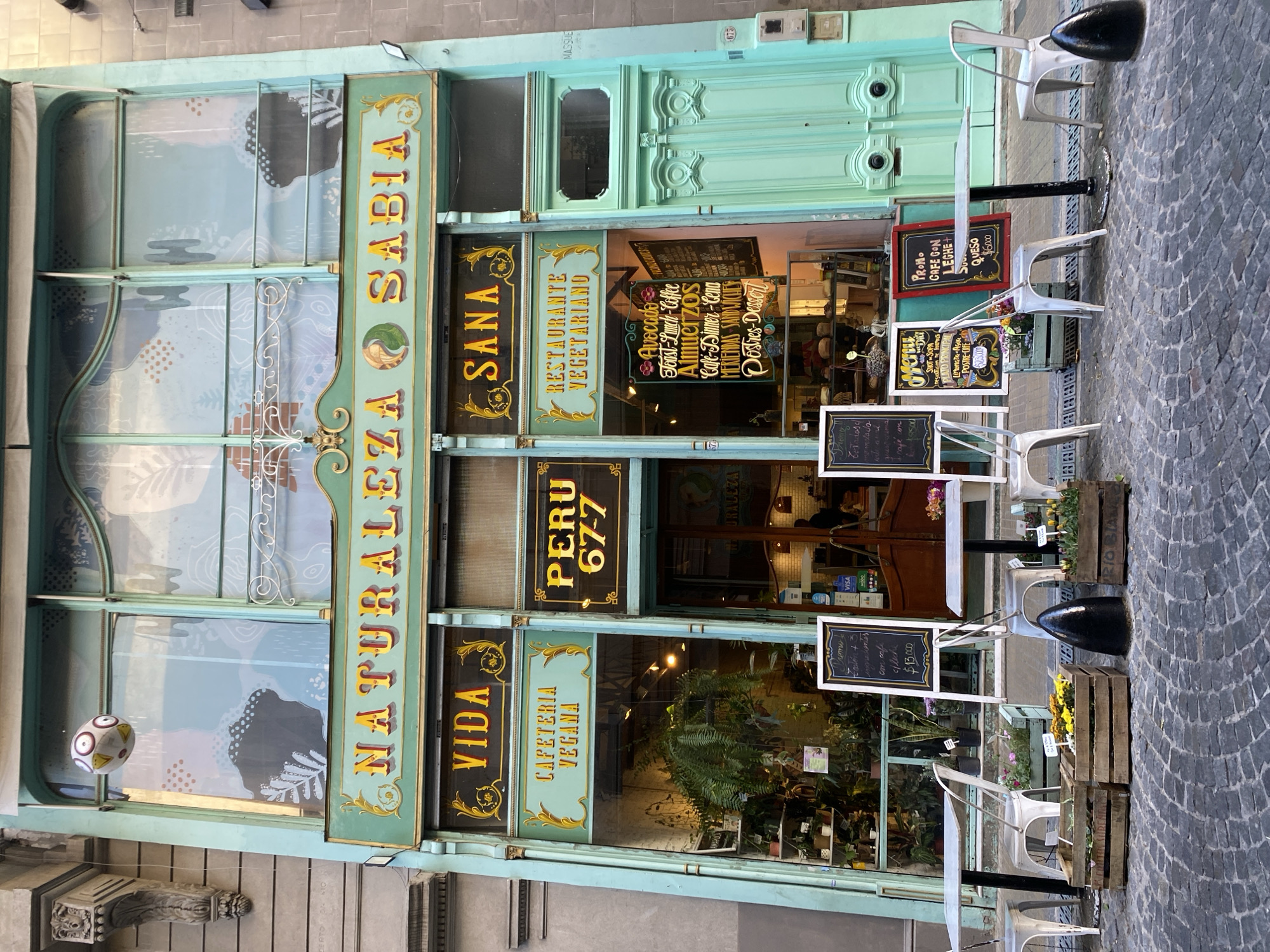 The image shows the front of "Naturaleza Sabia", a vegetarian restaurant and vegan cafe located at Peru 67-7 in Buenos Aires, Argentina. The building facade is painted in a light green color with gold accents on the signs. The establishment has large glass windows and a door with a transom window above. Signs advertise "Restaurante Vegetariano", "Cafeteria Vegana", and the address. Several blackboards display menu items and promotions, including "Avocado Toast-Lunch-Coffee-Almuerzos", "Cafe-Dinner-Cena-Meriendas Sandwiches", and "Postres-Dessert". Outdoor seating is arranged with white chairs and small tables, interspersed with wooden crates containing plants and flowers. The street is paved with cobblestones. A decorative egg is suspended inside the glass pane of the window.