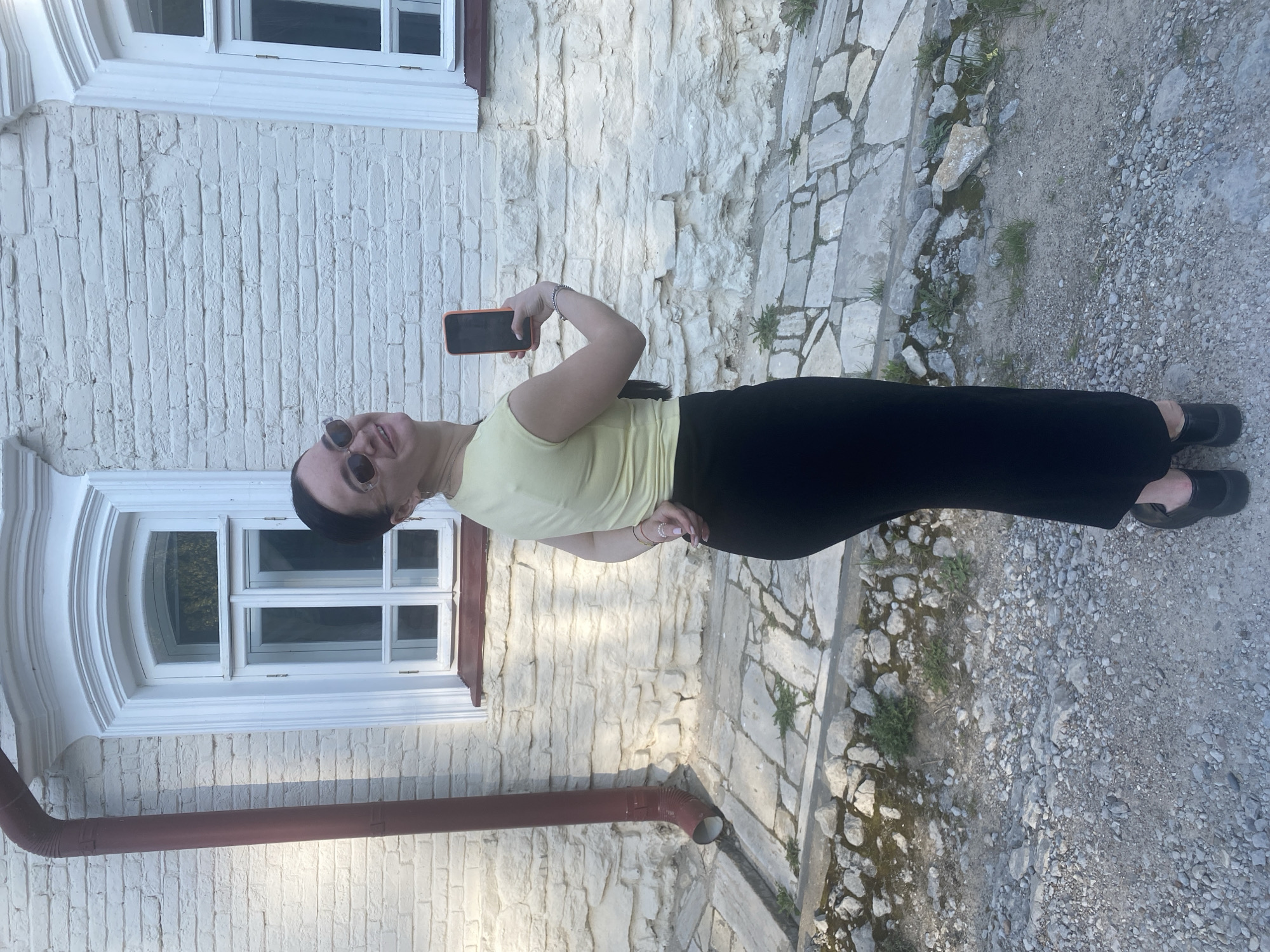 A woman with short dark hair, wearing sunglasses, a pale yellow tank top, and black pants, is taking a selfie with an orange smartphone. She is standing beside a white, brick building with a window featuring an arched top and white trim. A red drainpipe runs down the side of the building. The ground is paved with stone and gravel. The location is likely in Sviyazhsk, Russia, based on the building style.