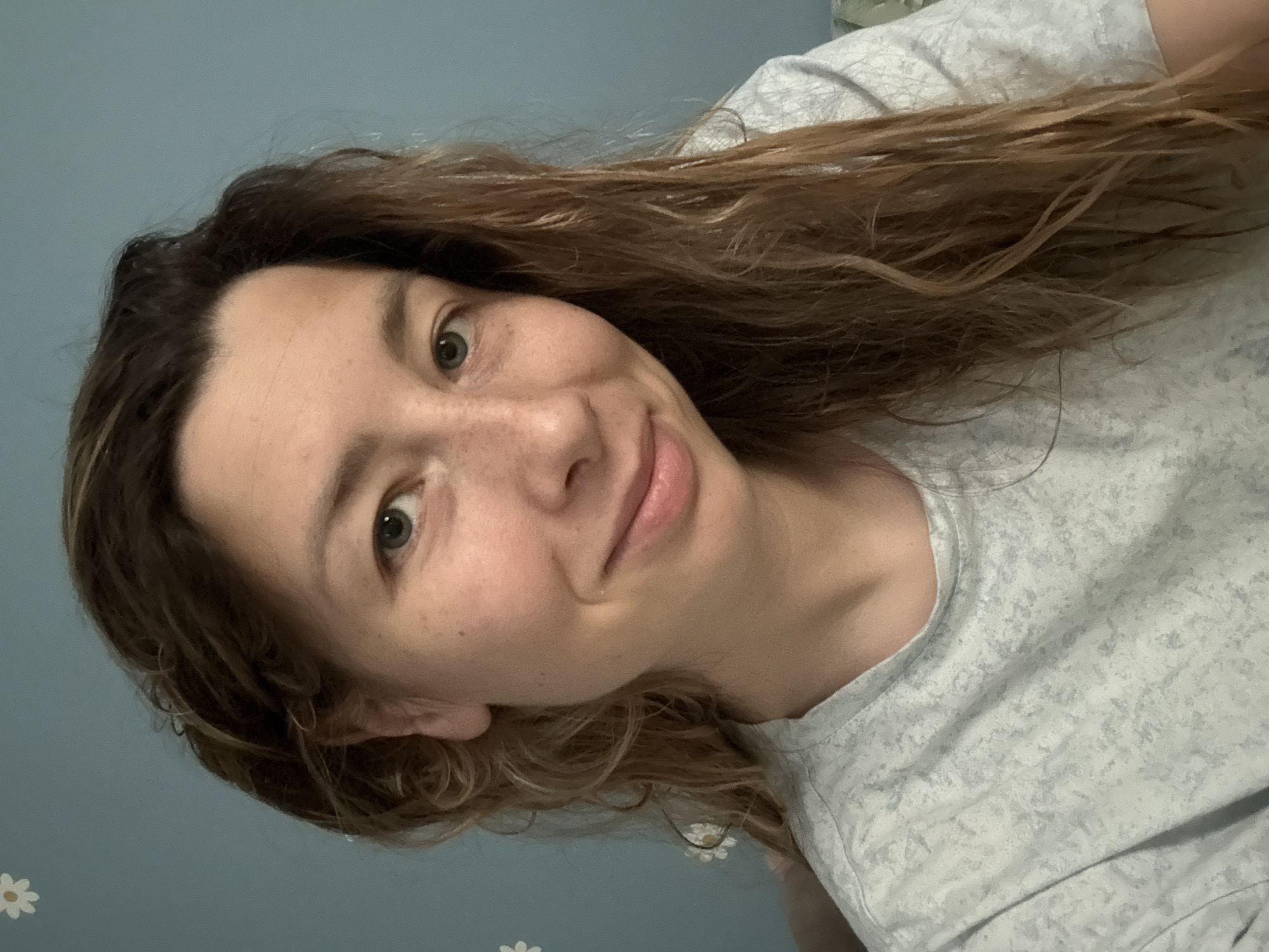 A medium shot photograph captures a woman with long, wavy brown hair and light skin with freckles, looking directly at the camera with a slight smile. She is wearing a light gray t-shirt with a subtle pattern. The background is a solid blue wall, with a small white daisy sticker visible in the upper left corner. The lighting appears natural, casting soft shadows. There are no discernible Moscow landmarks or references in the image.