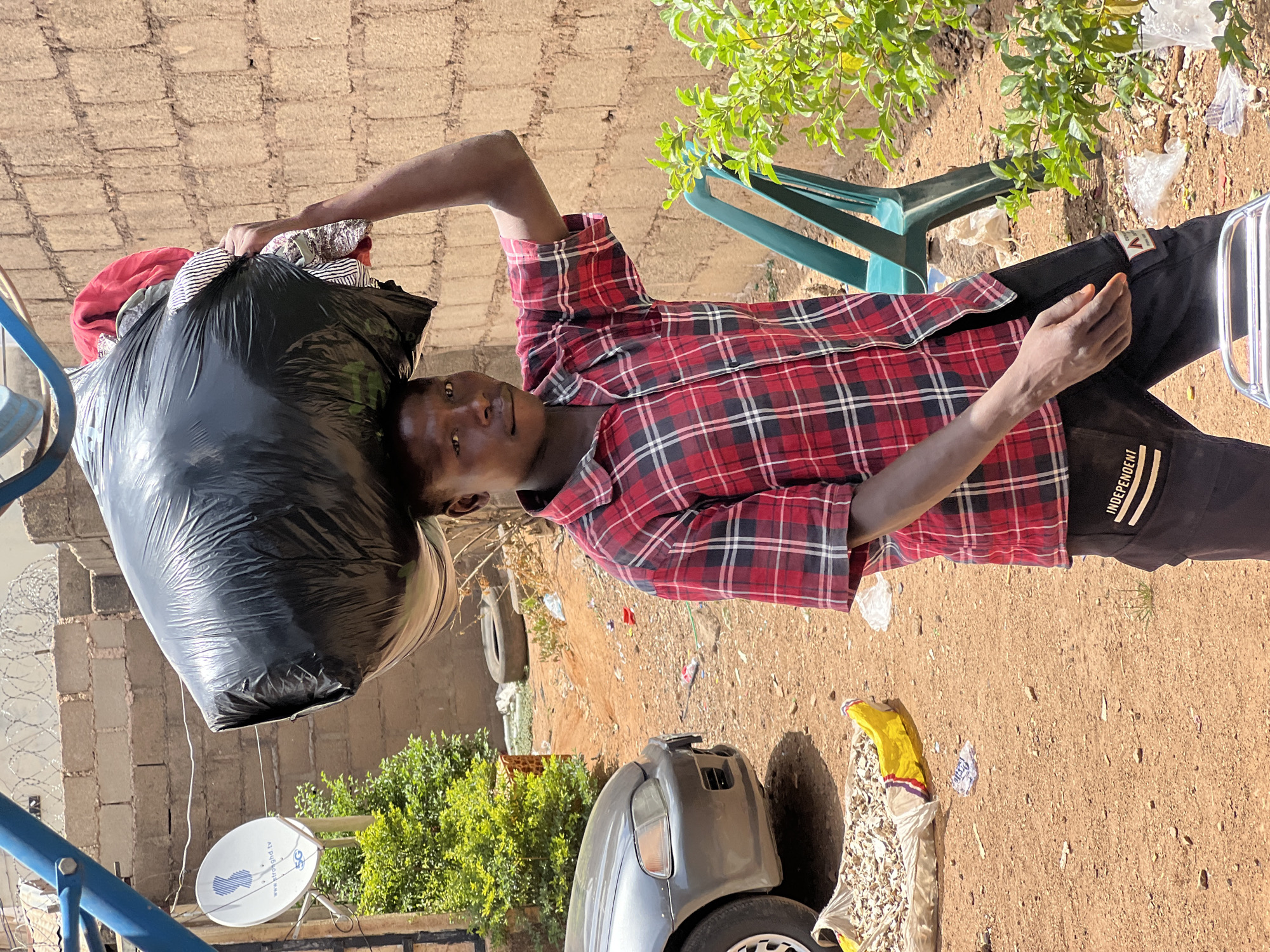 The image, oriented 90 degrees clockwise, depicts a young man standing outdoors in Jos, Nigeria. He is holding a large, full black plastic bag over his left shoulder, appearing to carry clothes or soft goods. The man wears a red, black, and white plaid long-sleeved shirt and black pants with a white stripe on the left leg. He looks directly at the camera with a neutral expression.
The scene is an unpaved, dusty outdoor area, likely a yard or informal space, under bright daylight. A wall made of light-colored cinder blocks, topped with barbed wire, is visible in the background. Green foliage from trees or shrubs is present on both sides of the frame.
Notable objects include a green plastic chair behind the man, a partial view of a gray car's front bumper and wheel, and a single worn yellow boot on the ground. On the far left, a blue metal structure is visible, along with a white satellite dish displaying "StrongHD.tv" and a "5G" logo. The ground is bare earth with scattered debris. Visible text includes "INDEPENDENT" on the man's pants and the branding on the satellite dish.
