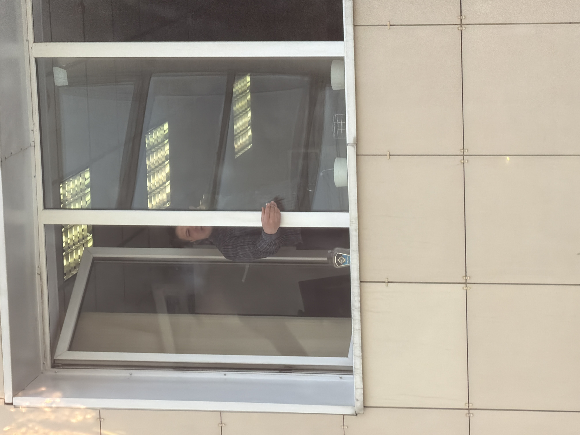 The image shows an open window in a building, likely a multi-story structure. A person is visible through the window, wearing a gray shirt. The window frame is white. Outside, the building has a tiled facade with light beige tiles. Reflections of what appear to be interior lights can be seen on the window glass. A Moscow city police emblem can be seen on the right side of the window. Given the emblem, the building may be a municipal building or a police station in Moscow, Russia.