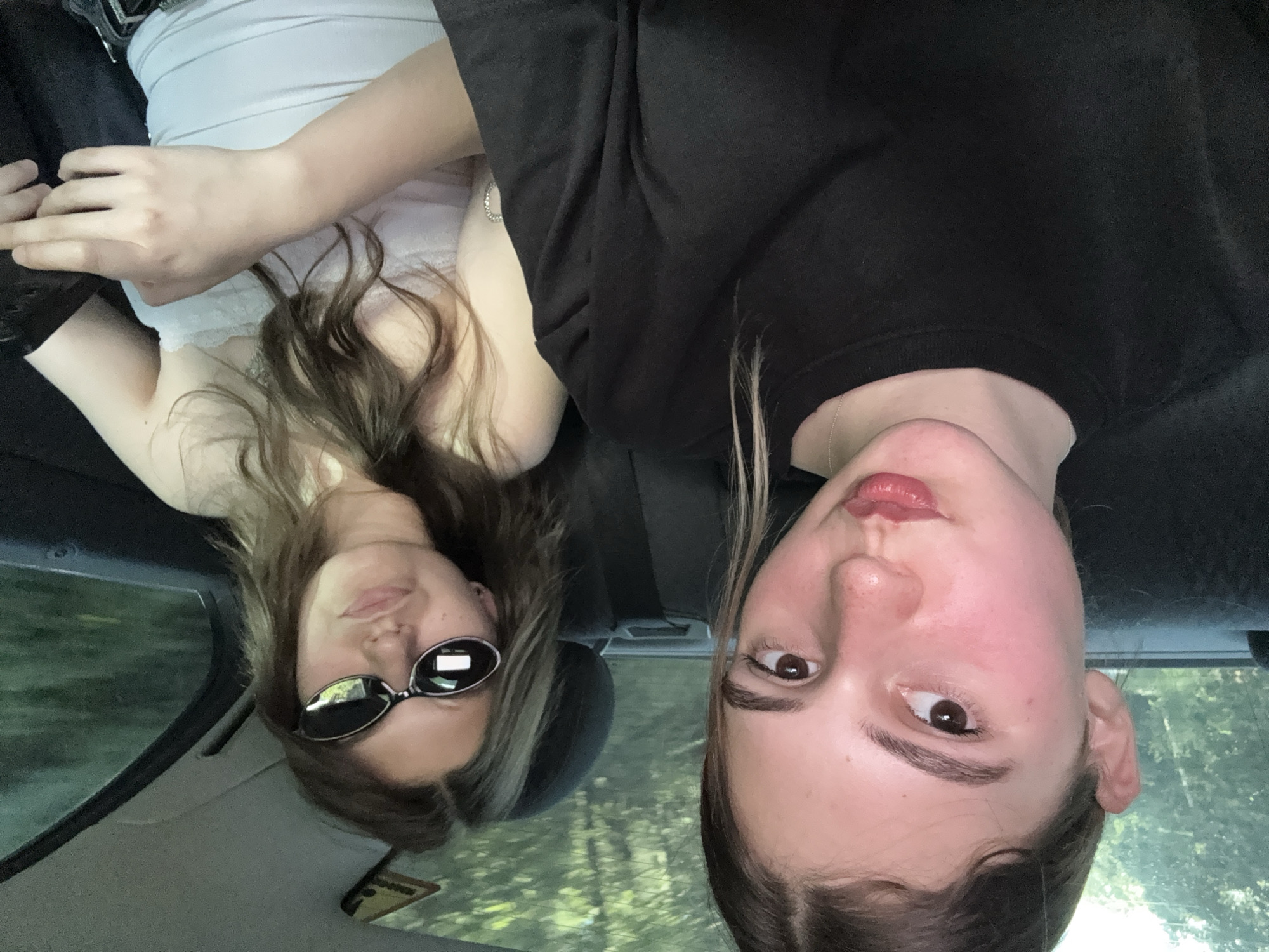 This is a slightly blurry, low-angle, upside-down selfie taken from inside a vehicle. Two young women are the primary subjects. The woman on the left is wearing a white tank top with lace trim and sunglasses. Her hair is brown and wavy. The woman on the right is wearing a black t-shirt. Her hair is dark brown, and she has rosy cheeks. The background appears to be a wooded area with green foliage, suggesting it's daytime and likely warm weather. The interior of the car is visible, with a gray headliner and a dark-colored seat or panel. The image conveys a casual and possibly spontaneous moment. There is no visible text in the image. The location is identified as Koprivshtitsa, Bulgaria.