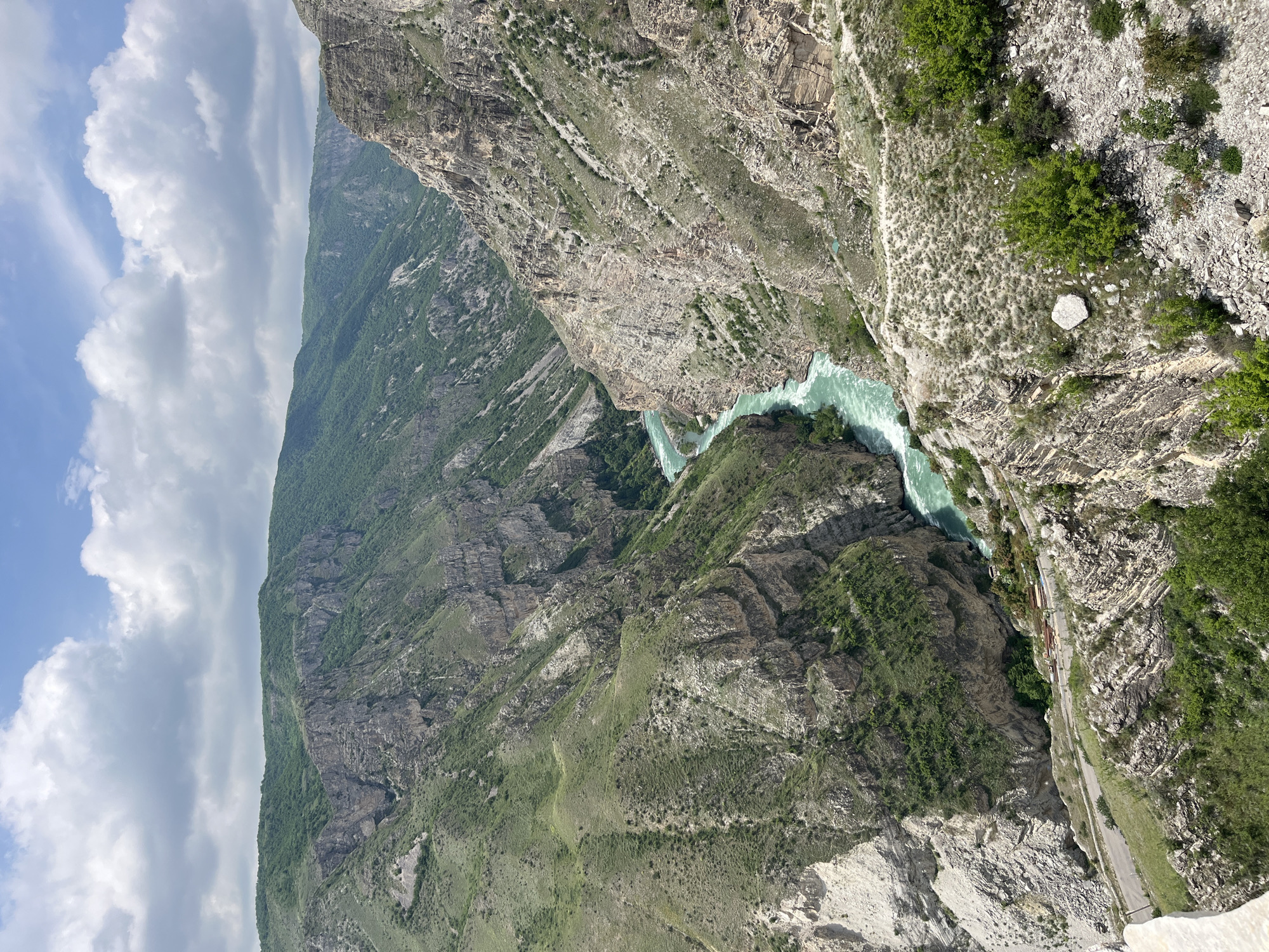 An aerial view of the Sulak Canyon near Chirkey, Russia. A steep-sided gorge contains a turquoise river. The canyon walls are made of layered rock formations with sparse vegetation. A road runs along the edge of the canyon on one side. The sky is partially cloudy.