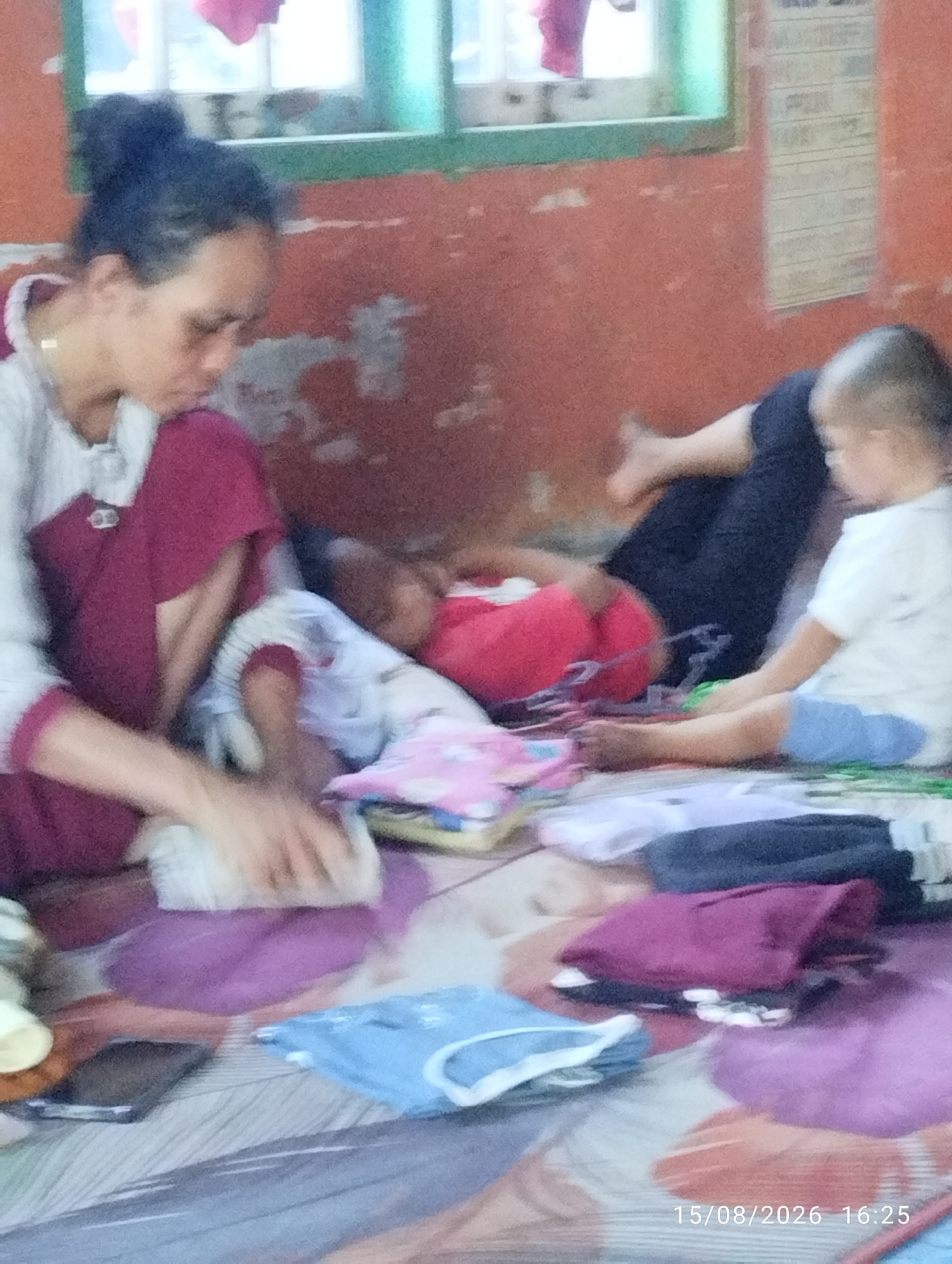 An indoor scene, likely a residential setting in Tegal Rejo, Indonesia, depicts three individuals on a floor covered with a patterned blanket. In the foreground left, a woman with dark hair tied up is seated, wearing a white shirt with a maroon overlay. She appears to be attending to a child. To her right, a young child is lying down, with their head resting on a red surface and their body partially covered by a light-colored garment. Further to the right, another young child is also lying down, wearing a white shirt and blue shorts, and appears to be interacting with items on the blanket. Various folded clothes in different colors, including purple, blue, and gray, are scattered around the individuals on the blanket. A dark rectangular object, possibly a mobile phone, is visible to the left of the woman. The background shows a wall with an orange-red hue and a window with green framing, suggesting a simple interior environment. A timestamp in the lower right corner reads "15/08/2026 16:25".