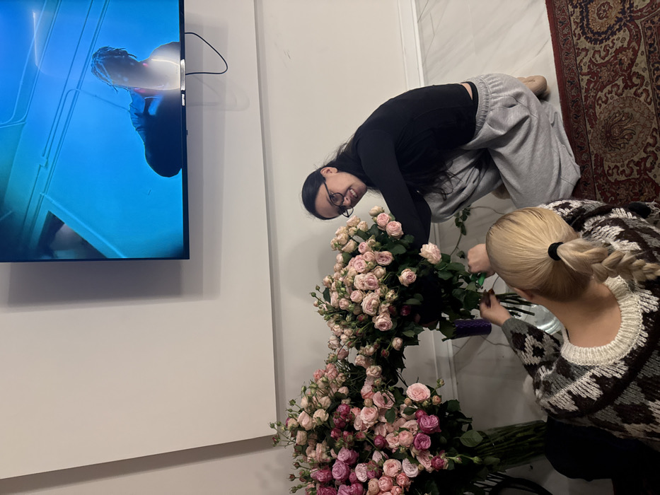 Two women arrange large bouquets of pink and light pink roses. One woman with dark hair and glasses kneels beside the flowers, smiling at the camera. The other, with blonde hair in a braid, focuses on trimming the stems. A TV hangs on the wall to the left, displaying a person in a blue setting. A patterned rug is visible in the upper right corner. The scene is set in a bright, modern room with a casual and comfortable vibe.