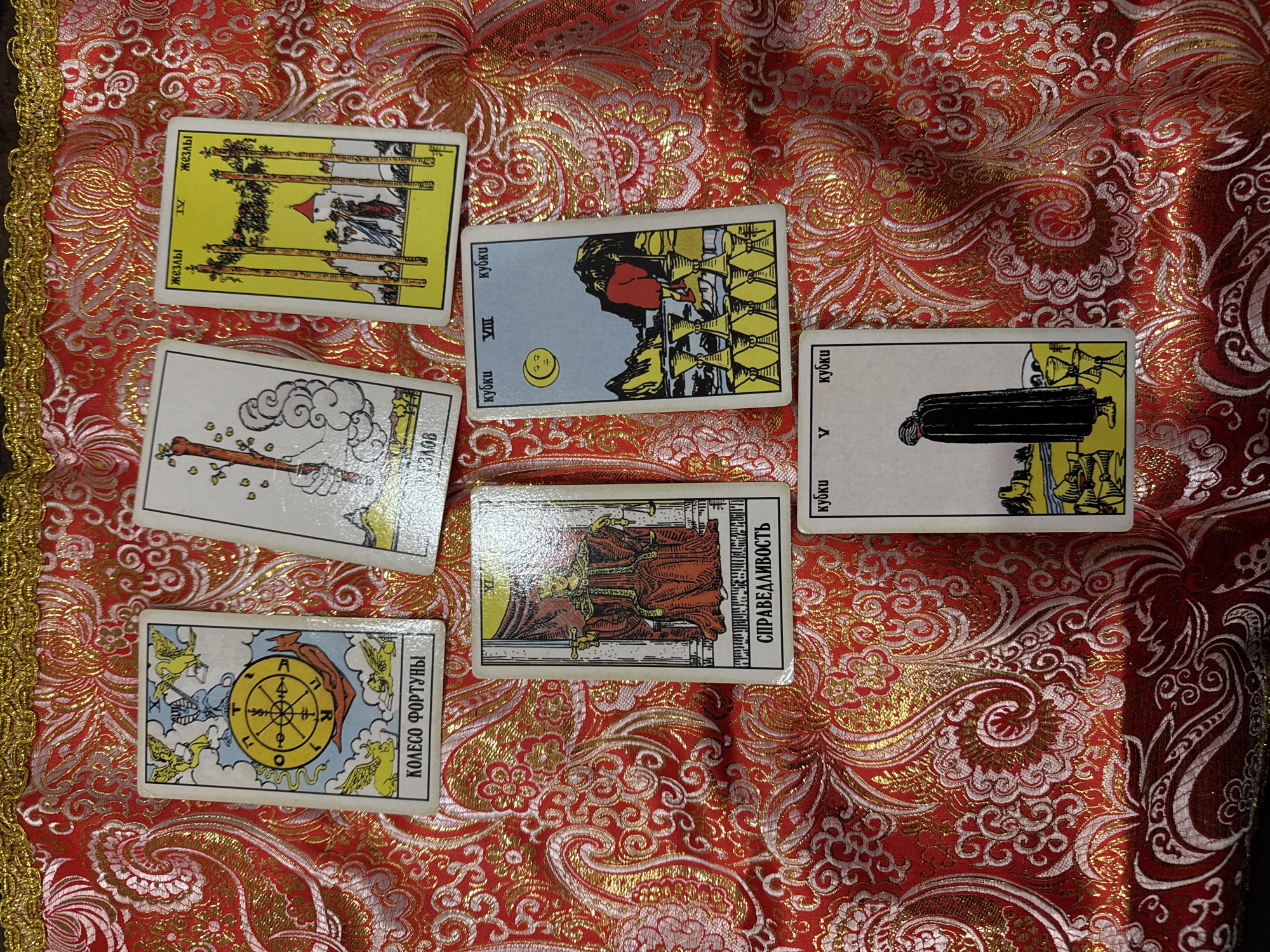The image shows a tarot card spread on a red and gold patterned cloth. The cards are laid out face-up and include, from top-left to bottom-right: Four of Wands, Eight of Cups, Five of Cups, Ace of Wands, Justice, and Wheel of Fortune. The Russian words for the card titles appear on the cards. There is no information in the image that indicates a specific location, such as the city of Sochi, Russia.