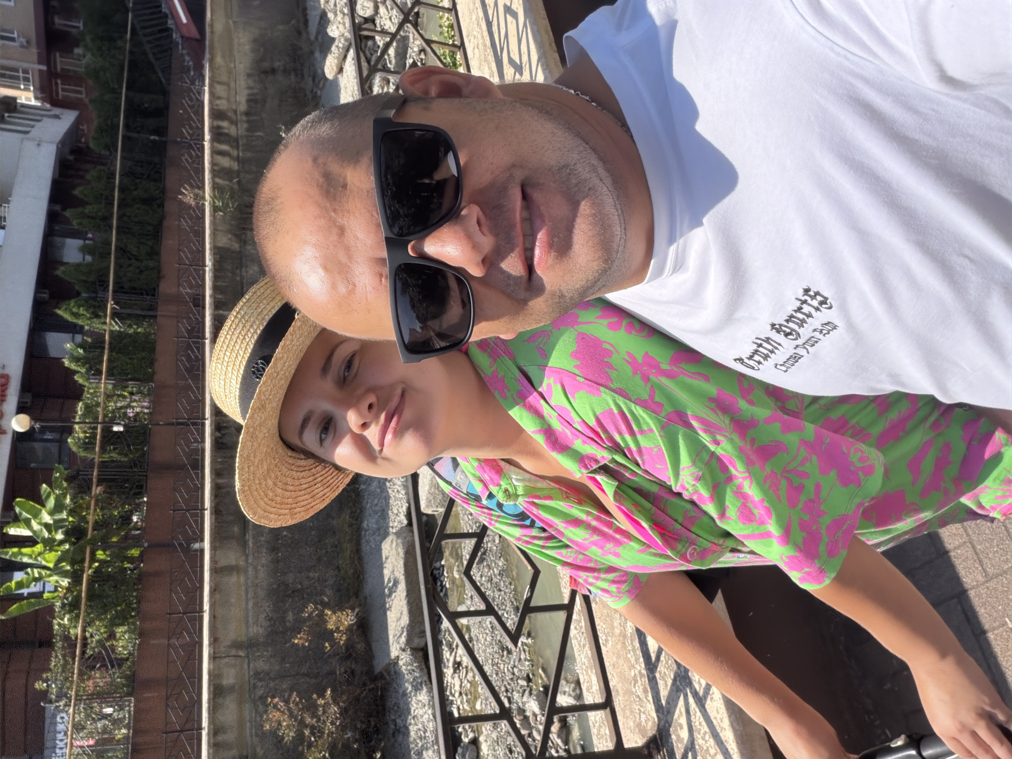 A man and a woman are taking a selfie outdoors in front of a river in Sochi, Russia. The man, in the foreground, is wearing a white t-shirt and black sunglasses, and he is smiling at the camera. The woman, to the left, is wearing a straw hat and a bright green and pink patterned shirt. She is also smiling. The river flows in the background, with a stone wall and a metal railing alongside it. Buildings and trees are visible in the distance. The lighting suggests it is daytime.