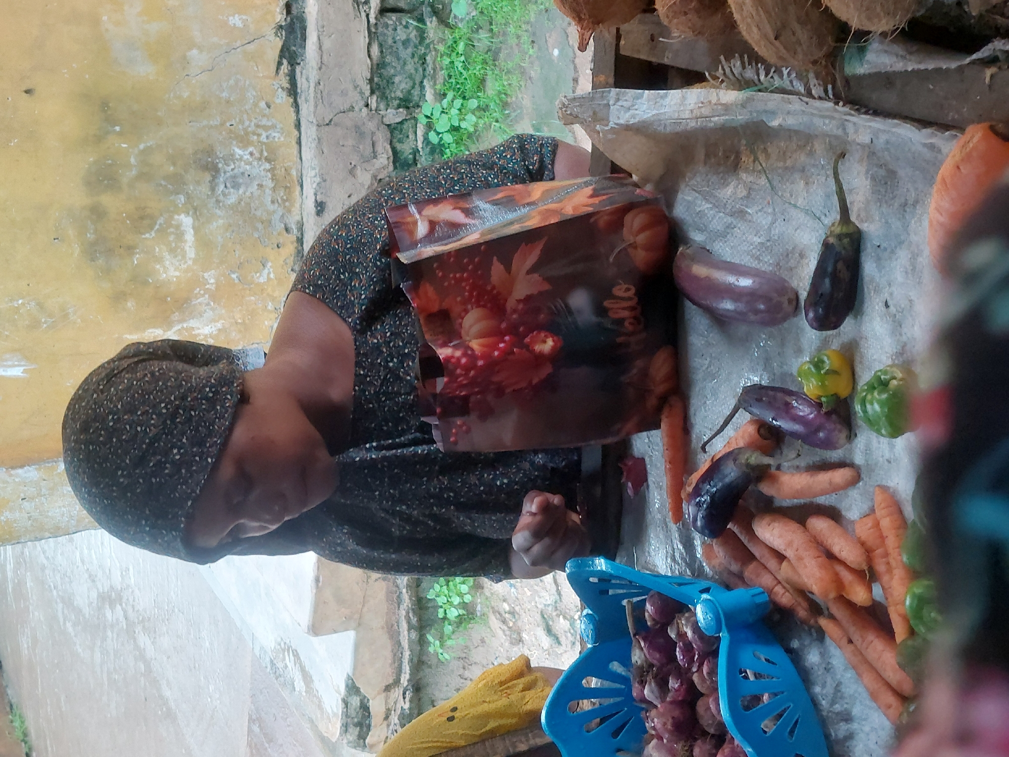 A woman in a headscarf and patterned dress is working at an outdoor market stall in Dar es-Salaam, Tanzania. She is positioned beside a display of vegetables, including carrots, eggplant, green peppers, and onions in a blue basket. A shopping bag with fall-themed imagery lies beside the produce. Coconuts are visible at the top of the frame. The setting is simple, with a yellow wall and steps in the background.