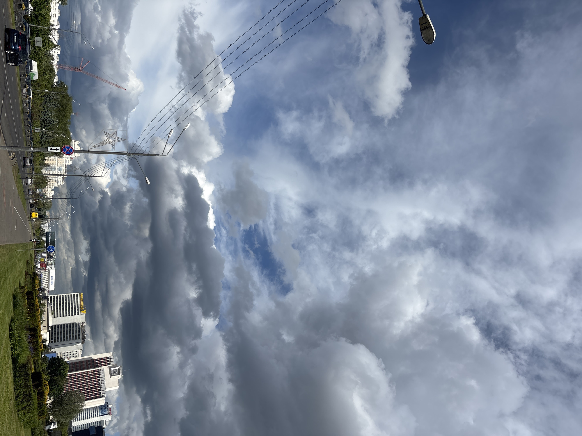 The image depicts an urban landscape in Minsk, Belarus, under a sky dominated by cumulonimbus clouds. Buildings are visible on the left side, including a hotel with the name "TROCIIHA" on the roof. A street runs alongside the buildings. A construction crane is present. Street lights and power lines extend across the frame. A road sign is visible along the street. The image is oriented vertically, with the sky occupying a significant portion of the frame.