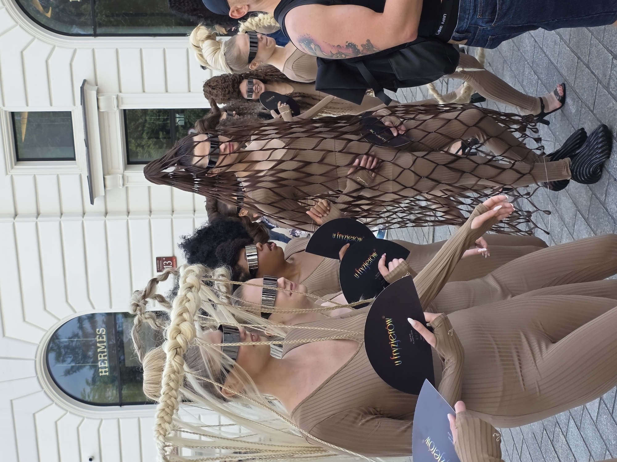 In front of the Hermès store in Warsaw, Poland, a group of people are participating in a fashion event. Several individuals, mostly women, are dressed in nude-colored, form-fitting outfits. They are adorned with elaborate braided hairstyles, some extending outwards in dramatic fashion. Many are wearing futuristic-looking sunglasses. Some are holding black fans with white text that reads "u fryzjerów". The overall aesthetic is avant-garde and theatrical. The street is paved with stone tiles, and the background features the white facade of the building with large arched windows, characteristic of the city's architecture.