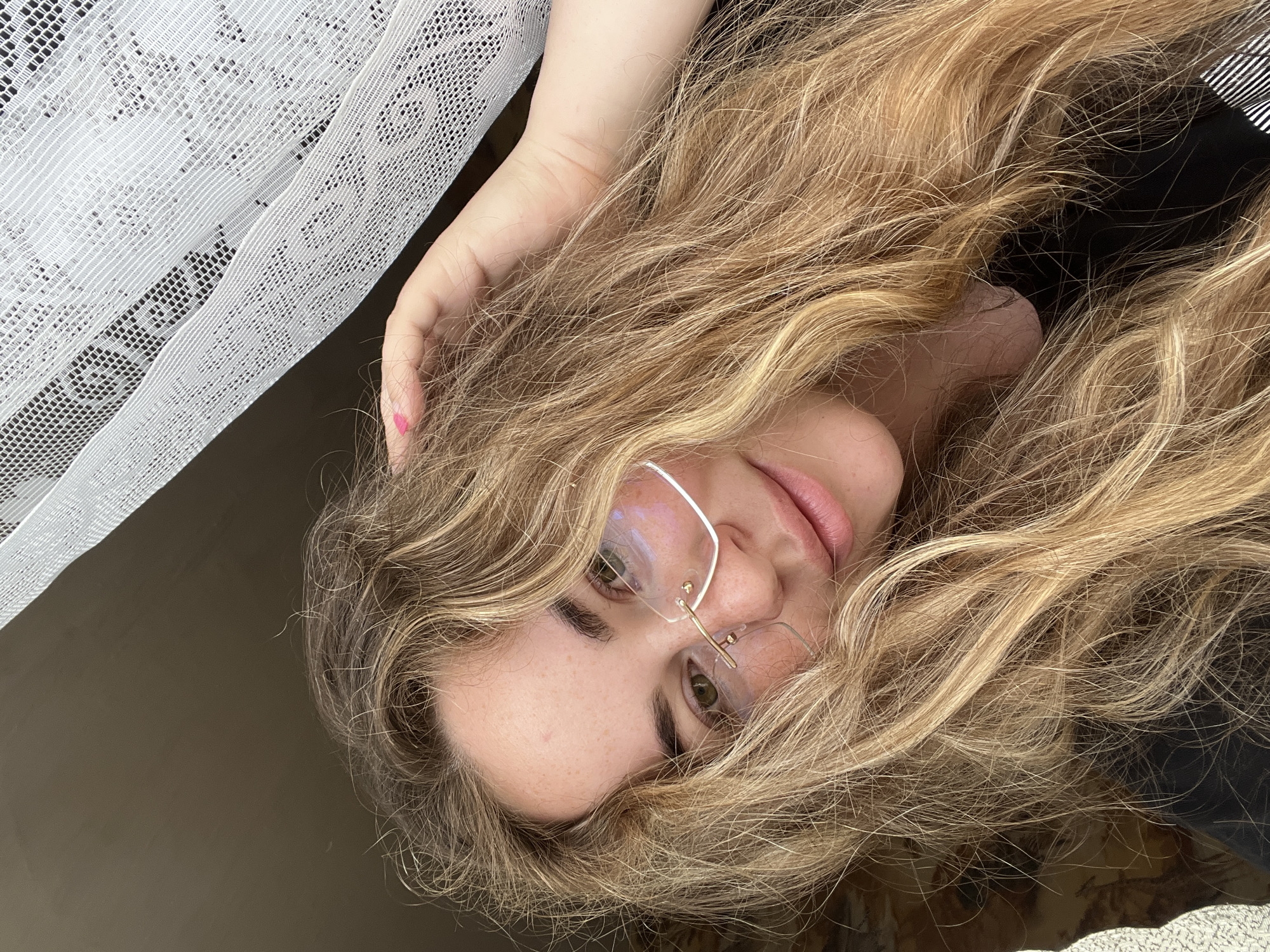 A close-up portrait depicts an individual, likely female, with long, wavy, light brown and blonde hair, partially obscuring the face. The subject wears rimless eyeglasses with gold-toned accents and has visible freckles on the forehead and nose. Green eyes are visible through the lenses, which show reflections of an external light source. The lips are slightly parted, displaying a natural pink color. The subject's left hand is positioned in the hair near the crown of the head, revealing a pink heart design on the nail of the left index finger. A dark-colored garment, possibly black, is visible around the neck and shoulder area, with a partial black and white striped pattern visible on the subject's right side, consistent with a collar or strap. The background to the upper right features a white, translucent lace curtain with an intricate patterned mesh, indicating a window. A dark, uniform surface is visible behind the upper left portion of the head, and a lighter, textured fabric is partially visible in the lower left quadrant of the image. The scene is situated in Сосново, Russia.