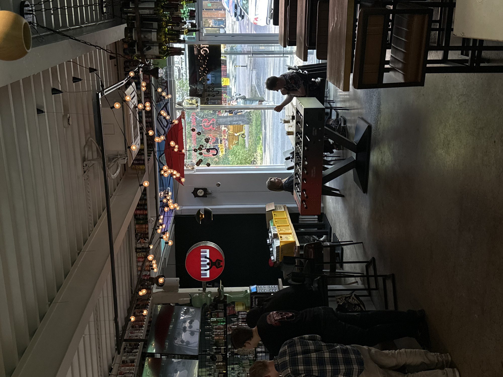 The image depicts the interior of a bar in Khimki, Russia. In the foreground, two men are standing at a bar counter. One man is wearing a plaid shirt and light-colored pants, while the other is wearing a black t-shirt with a graphic print and dark pants. Behind them, more people are visible at the bar, and the bar is stocked with bottles. Further into the bar, a foosball table is set up, with two individuals seated on opposite sides. One person, wearing a gray top and dark pants, is seated on the left, facing the foosball table. On the right, a person in a dark patterned shirt is leaning forward and appears to be playing foosball. The environment is dimly lit with string lights and overhead lighting. A red umbrella-shaped decoration hangs from the ceiling, adorned with lights. Large windows in the background offer a view of the street outside, where cars and pedestrians can be seen. The floor is concrete, and several tables and chairs are arranged throughout the space. A circular sign with a piano keyboard and a silhouette of a person is mounted on the wall.