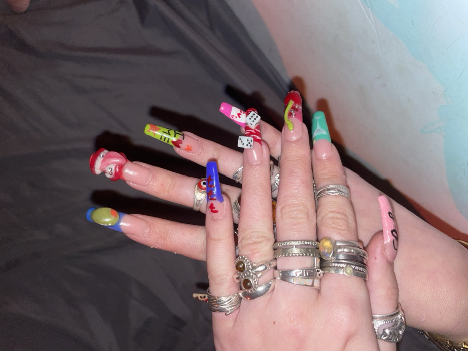 A pair of hands adorned with long, sculpted nails and numerous rings is displayed. Each nail features a unique and elaborate design, including dice, a pair of lips, food items and vibrant colors. The rings vary in style, some stacked and others featuring gemstones. The hands rest against a backdrop of dark fabric and a lighter colored wall.