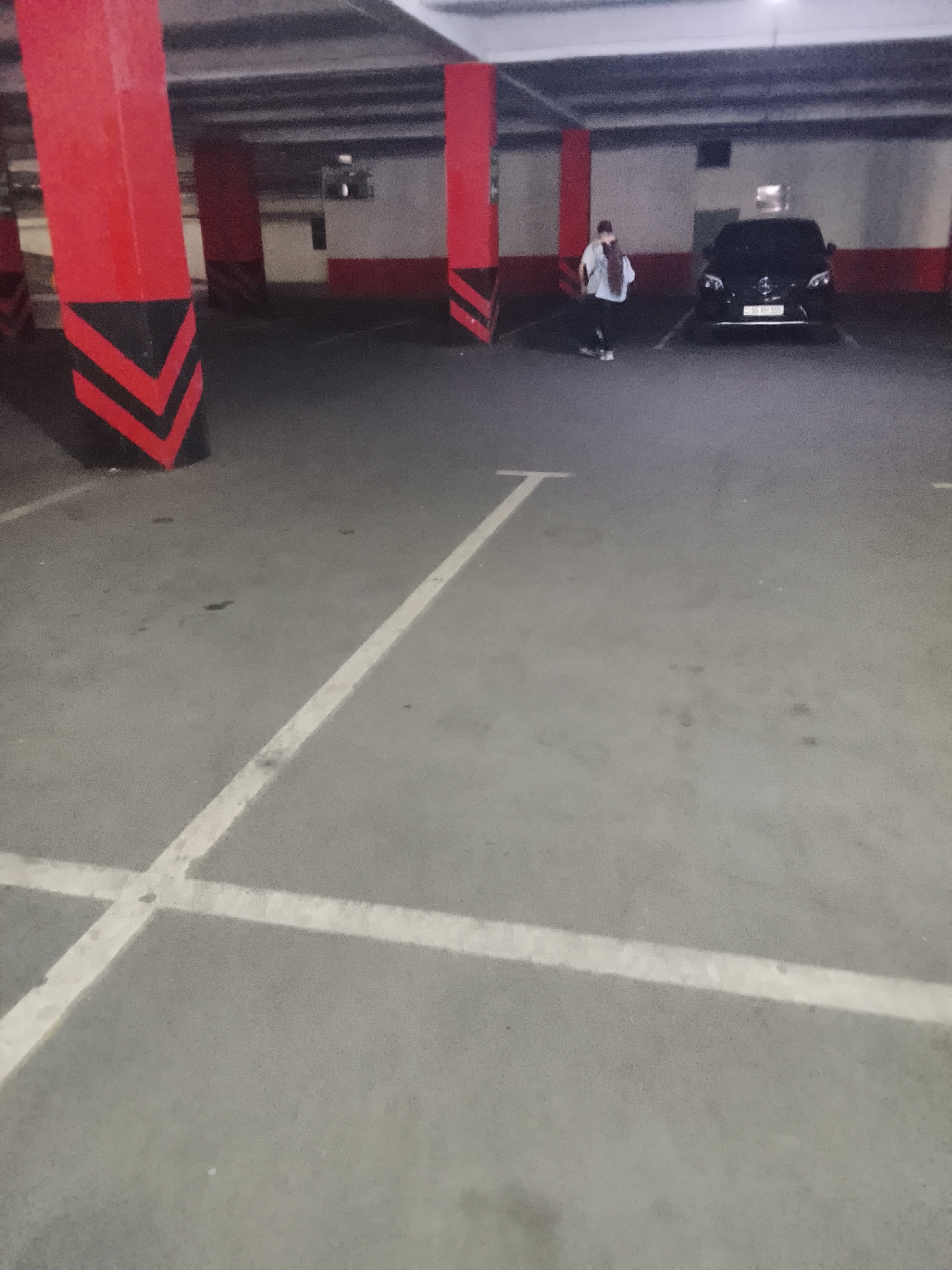 In an underground parking garage, a person wearing a white shirt, dark pants, and a backpack walks towards a parked black Mercedes-Benz sedan. The person is positioned to the left of the vehicle, with their back to the camera. The garage has red and black striped columns and white painted lines on the concrete floor, indicating parking spaces. The lighting is dim, typical of an indoor parking facility.