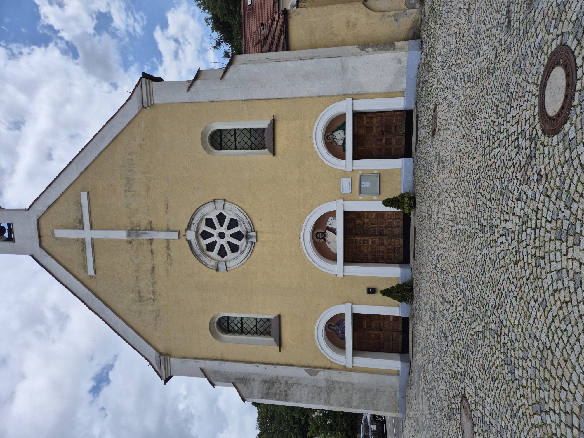 The image shows the facade of a yellow church in Landshut, Germany, on a cloudy day. The church has a large white cross on its upper facade, a circular rose window above the entrance, and two arched windows. The main entrance is divided into three arched doorways, each with a pair of wooden doors. Above each doorway is a religious mural. Two small evergreen trees are planted in front of the doors. The ground in front of the church is paved with cobblestones. A manhole cover is visible in the foreground. To the left of the church, a car is parked. The background shows trees and a reddish-tiled roof.
