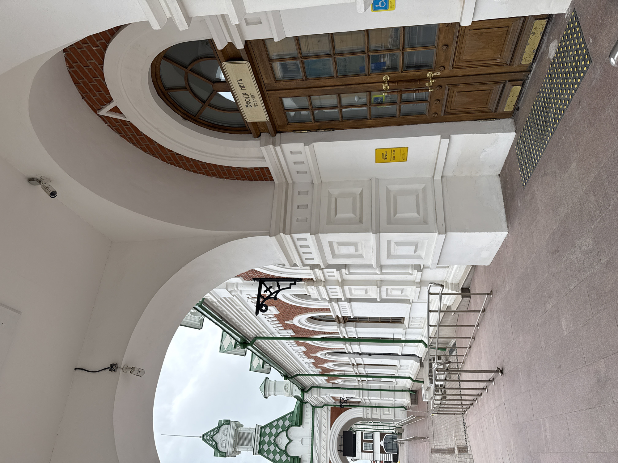 The image depicts a building entrance in Perm, Russia. The entrance features a pair of brown wooden doors with multiple glass panels, a circular window above the door, and a sign with the words "ВХОДА НЕТ" (No Entry). The architectural style includes white arches, decorative brickwork, and white rectangular patterns on the walls. A tactile paving surface is present in front of the door, and a handrail extends along the exterior. A surveillance camera is mounted on one of the arches. The weather appears overcast.