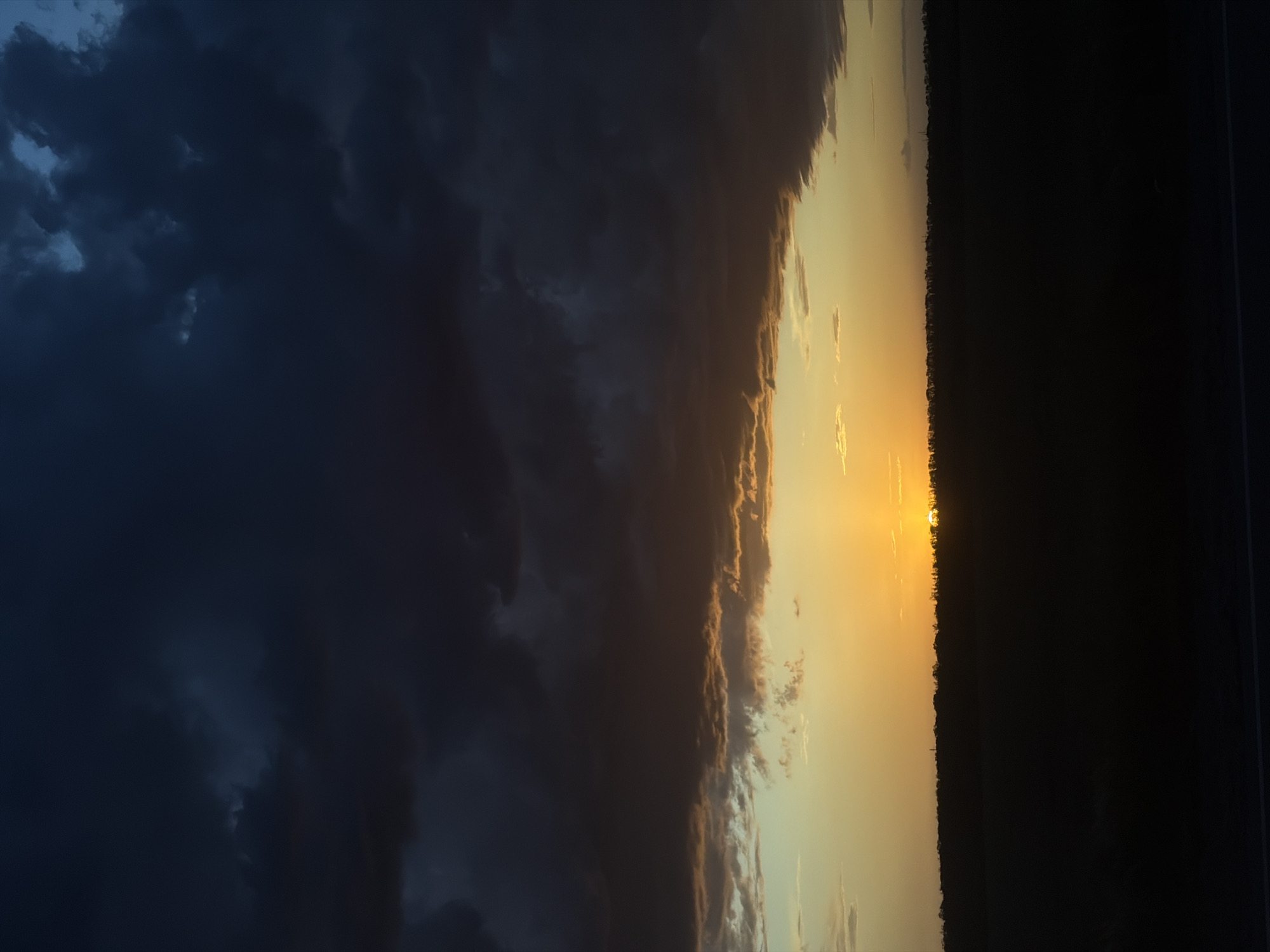 The image depicts a sunset scene, likely taken outdoors at dusk or dawn in an unknown location. The sun is obscured by the horizon, producing a bright orange glow illuminating the sky and silhouetting the landscape. To the left, a dark, cloudy sky contrasts sharply with the vibrant colors near the horizon. A dark, dense line suggests a treeline or distant landscape feature along the horizon. A road is barely visible on the right side.