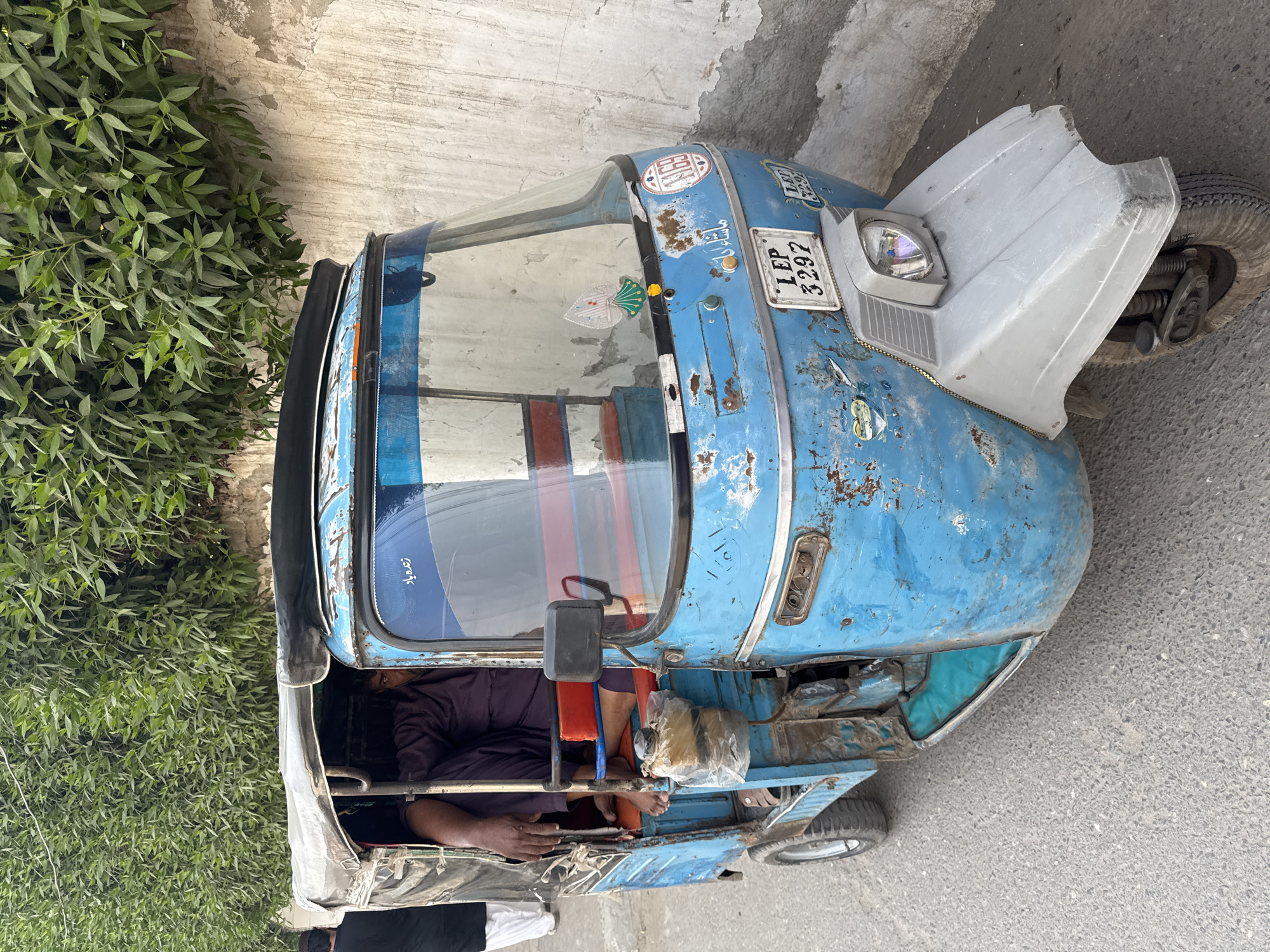 An auto-rickshaw, with license plate LEP 3297, is parked on a paved surface near a wall and green foliage. The auto-rickshaw is predominantly blue with visible rust and wear. The front hood is a light gray color. The vehicle’s interior shows a person in a dark purple shirt. The setting is Ghareeb Abad, Pakistan.