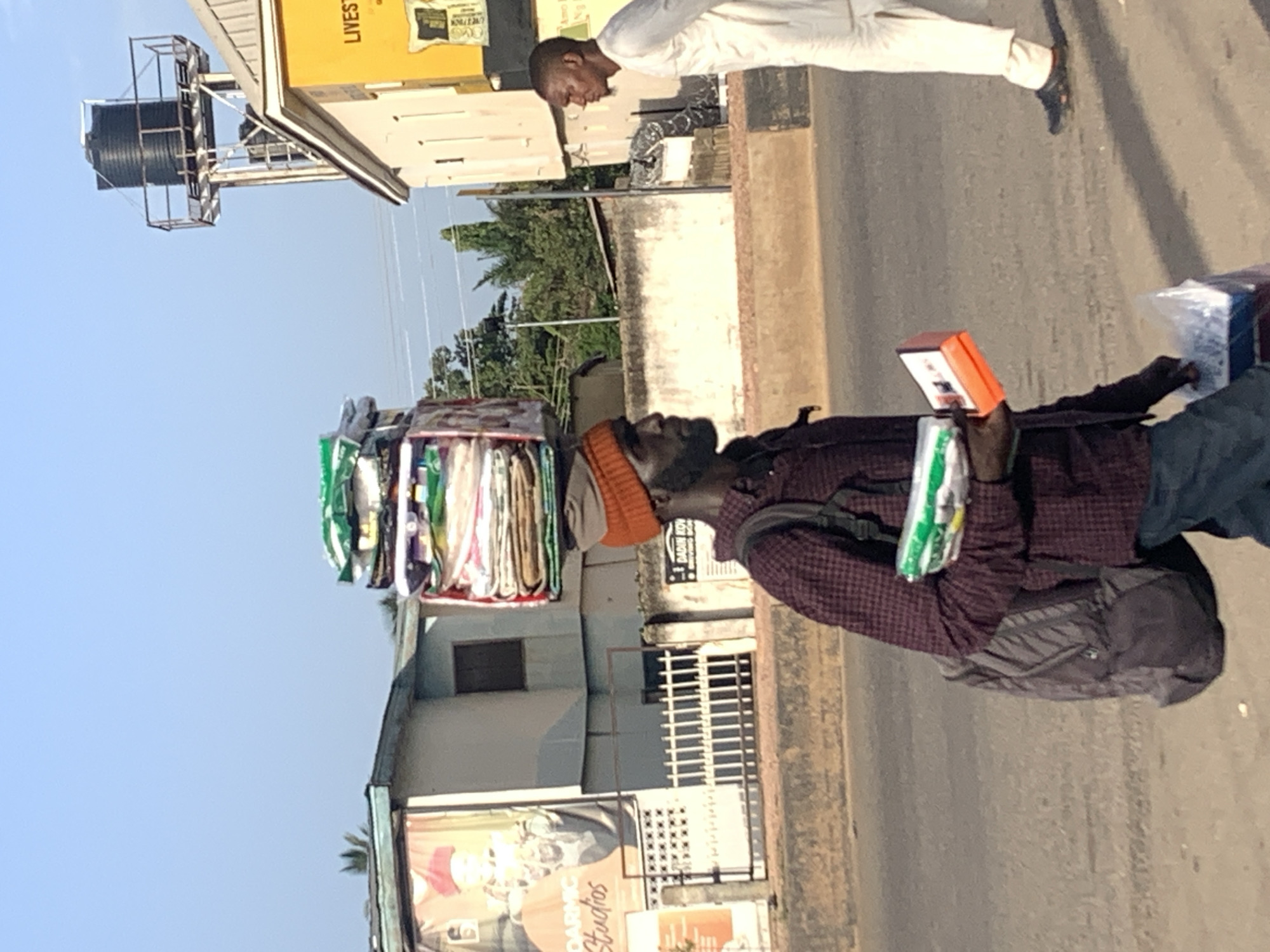 A man with dark skin and a beard, wearing a maroon checkered shirt and an orange beanie, walks along a road in Jos, Nigeria. He carries a large stack of items balanced on his head, wrapped in plastic and fabric. In his right hand, he holds a green and white package and an orange box. He also has a grey backpack slung over his left shoulder. Another man, with lighter skin, wearing a white robe and dark shoes, walks in the opposite direction. Behind the first man, a building with a large advertisement featuring a man in a red hat and the words "DARMIC Studios" is visible. Further in the background, a water tower stands tall. The environment appears to be urban, with buildings and a paved road.
