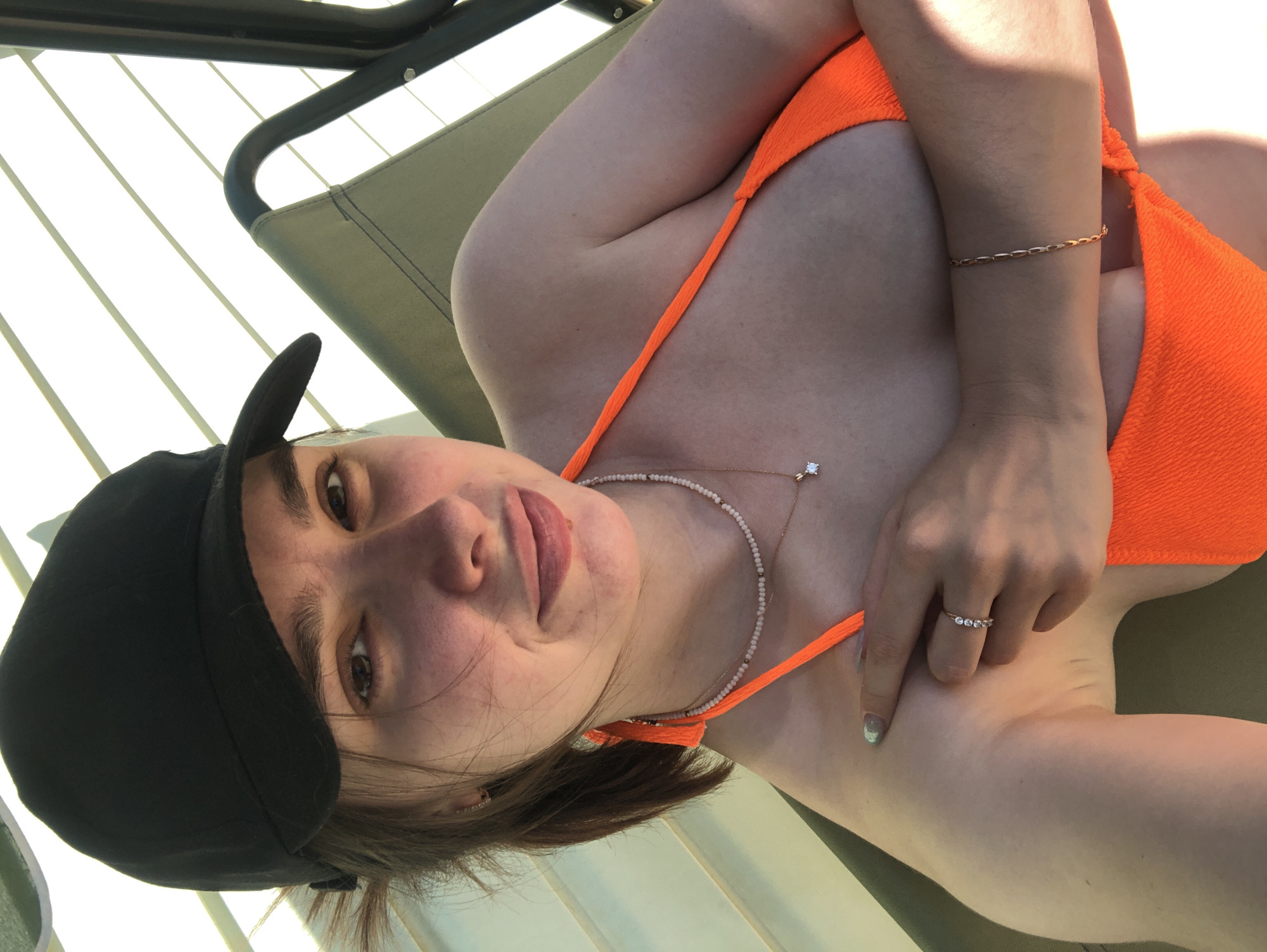 A woman wearing a black baseball cap and an orange bathing suit is lying on a chaise lounge. She is also wearing two necklaces and rings on her fingers. The location of the scene is not specified. The city of Noviy, Russia is not mentioned in the image.
