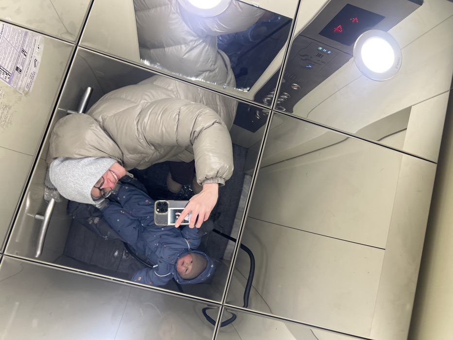 The image captures an adult and an infant reflected in a mirrored elevator interior located in Khimki, Russia. The adult, likely female, is centered in the reflection, wearing a light beige or taupe puffy winter coat over a grey hooded sweatshirt and black-framed glasses. She is smiling and holding a smartphone with a clear case in her right hand, actively taking the photograph. A grey knitted glove is visible on her left hand. To her left, an infant, wearing a blue patterned winter suit with a hood and a light brown knitted hat, is visible from the chest up, looking directly towards the camera. The infant appears to be seated in a dark, obscured carrier or stroller. The elevator walls and ceiling are covered with multiple reflective, silver-toned panels, forming a grid pattern. In the lower right portion of the reflection, an elevator control panel is visible, featuring various buttons, partially visible Cyrillic text, and a red digital display showing "2" and an upward arrow. A circular, illuminated light fixture is positioned below the control panel.