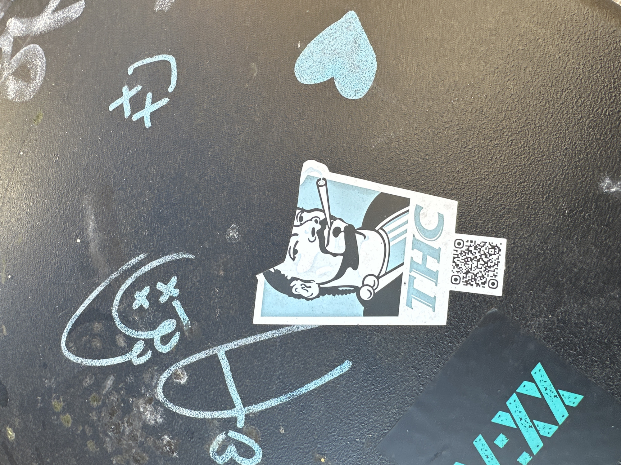 This image shows a close-up of a dark, textured surface, likely an outdoor table or a similar urban fixture, in London, United Kingdom. The surface is covered with various forms of graffiti and stickers. There are abstract chalk markings in light blue, including symbols resembling eyes and a heart. A sticker featuring a cartoon character smoking a cigarette with the letters "THC" and a QR code next to it is attached to the surface. A portion of another sticker with a design in teal and black is visible in the lower right corner. The environment appears to be urban, based on the nature of the surface and the graffiti. There are no people visible in the image.