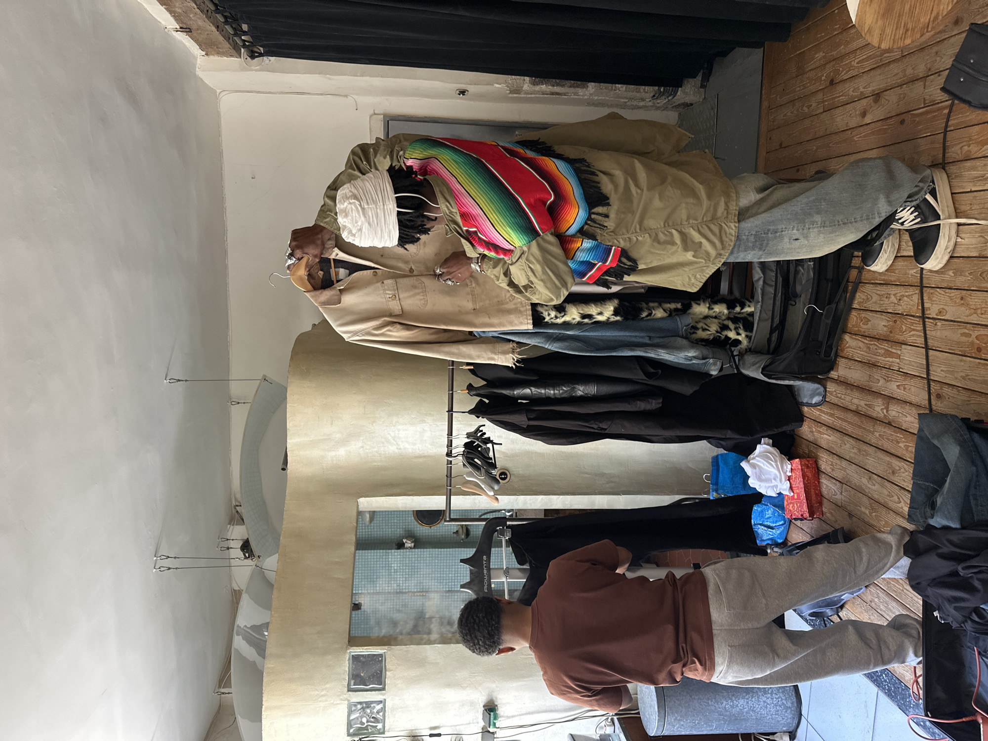 The image depicts the interior of a retail space, potentially a clothing store or boutique. A light-skinned person wearing a white turban, sunglasses, and a colorful striped garment is holding up two coats on hangers. One coat is a khaki color, and the other is a light brown color. Hanging racks filled with dark clothing, including what appear to be leather jackets, are visible. There are also piles of denim and other clothing items on the floor. In the foreground, another person wearing a brown shirt and grey sweatpants is operating what appears to be a clothes steamer. The setting includes a wooden floor and what appears to be tiled flooring in the background. Through a window, the blue walls of a bathroom or utility room can be seen. The architectural style suggests a European, possibly Parisian setting, due to the style of the window and general building design.