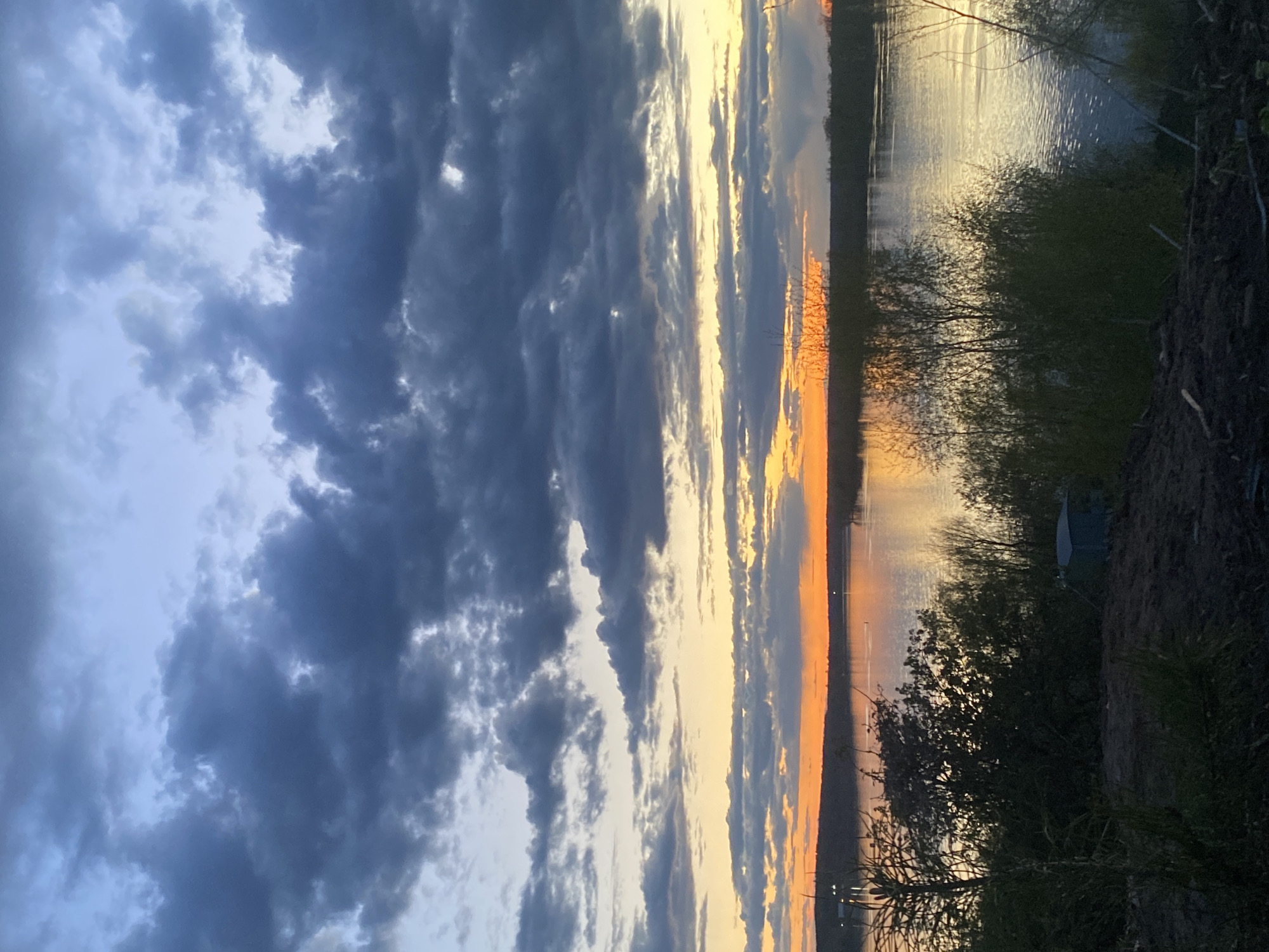 The image shows a sunset over a body of water, likely a lake or river, in or near Ольховка, Russia. Heavy, dark storm clouds dominate the left side of the frame. Light from the setting sun breaks through gaps in the clouds, creating bands of orange and yellow light in the sky and reflecting on the water's surface. A dark shoreline with vegetation is visible on the right side of the frame. A small structure, possibly a tent or shelter, is vaguely discernible near the shore. The image appears to be taken at ground level.