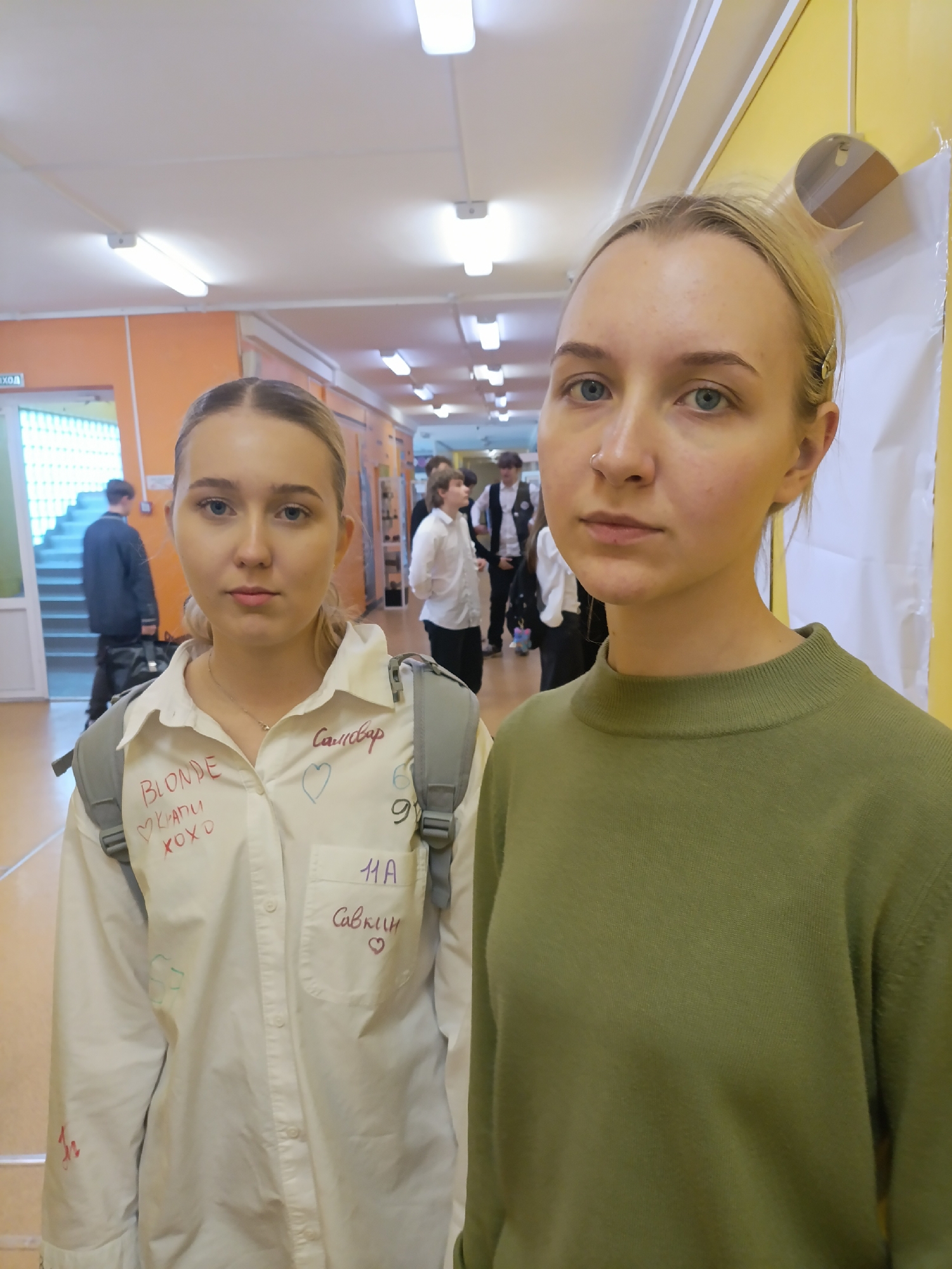 Two fair-skinned, blonde young women stand side-by-side in a brightly lit hallway with fluorescent lights. The woman on the left wears a white button-down shirt with writing in various colors, including "BLONDE," "XOXO," and names, along with hearts. She also has a gray backpack on. The woman on the right wears a plain olive green sweater. In the background, the hallway extends with other people visible, some in school uniforms. The walls are a mix of orange and yellow. There's a doorway visible in the background, along with various items hung on the walls. A sign above the doorway reads "ВЫХОД." Based on this sign, the location is determined to be in Krasnoyarsk, Russia.