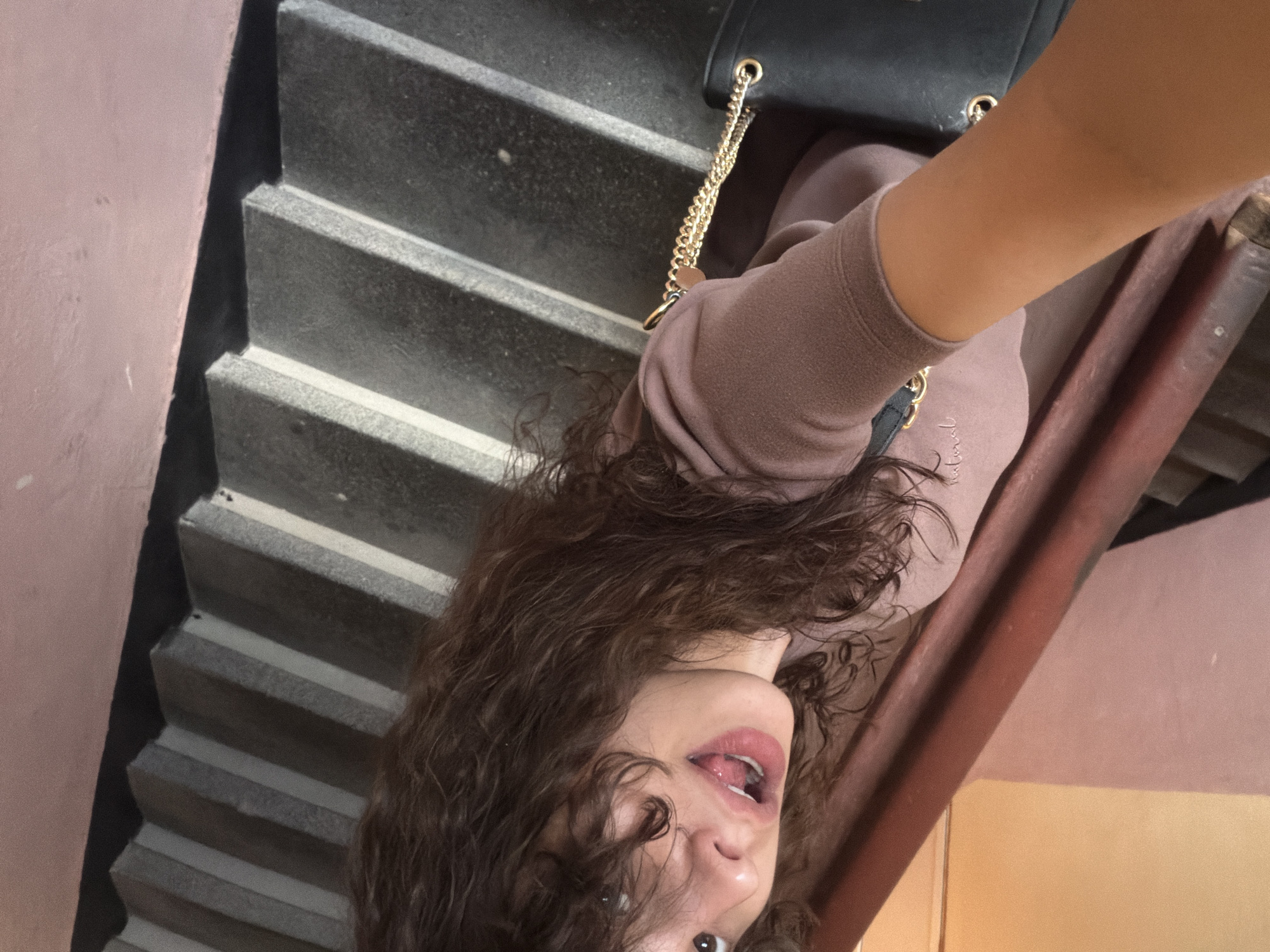 In this close-up, high-angle shot taken from the perspective of the subject, a young woman with curly brown hair is looking at the camera. She is wearing a mauve long-sleeved top and holding a black purse with a gold chain strap. Her mouth is slightly open, with her tongue visible. The background consists of a pink-painted wall and a set of concrete stairs with grey and white risers. The scene appears to be an indoor stairwell.
