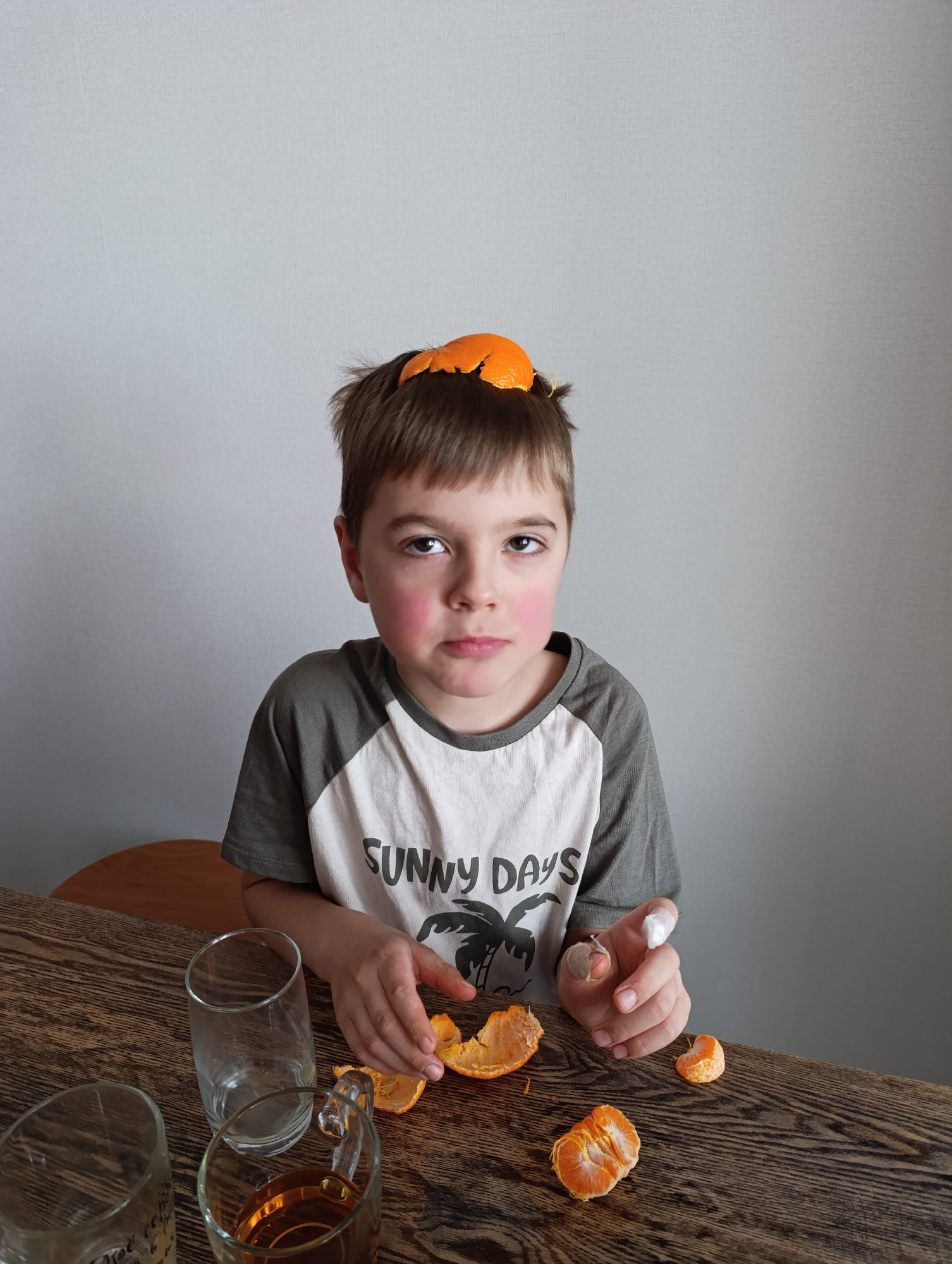 A young boy sits at a wooden table with pieces of mandarin orange peel scattered around him. A piece of the peel rests on top of his head. He wears a t-shirt with the words "SUNNY DAYS" printed on it. On the table are three glasses, one clear and empty, one with a yellowish liquid, and another partially visible with writing on it. The background is a plain light gray wall. The location is not specified in the image but the question states that it is Perm, Russia.