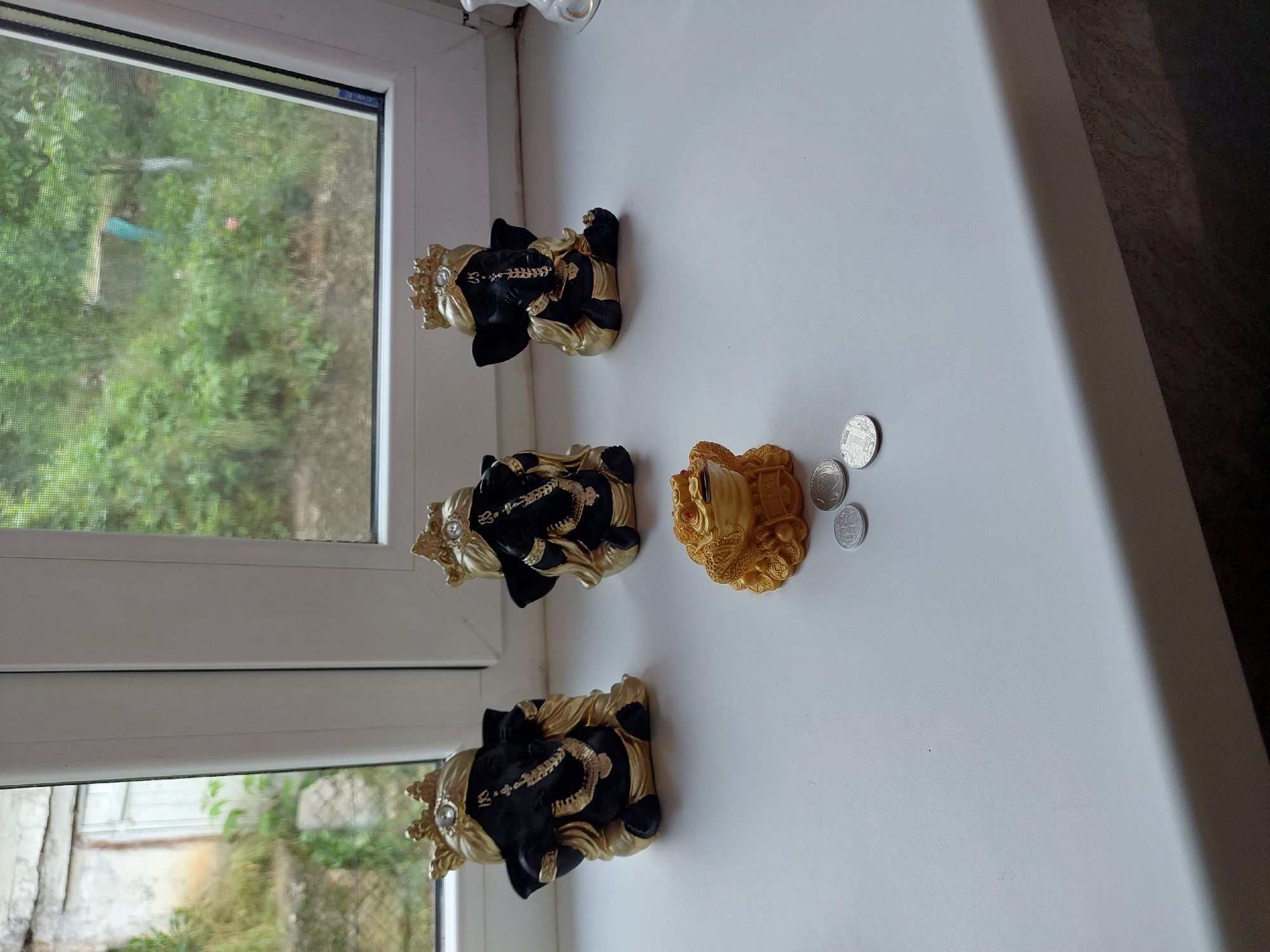 The image captures an interior view looking out through a white-framed window onto an exterior environment in Orhei, Moldova.
Occupying the white, matte-finished windowsill are three small statues of the Hindu deity Ganesha, arranged in a linear fashion from left to right. Each Ganesha figure is predominantly black with gold accents, including elaborate headwear (crowns featuring a central clear gemstone), wraps, and jewelry. From left to right, the Ganeshas appear to embody the "hear no evil, see no evil, speak no evil" motif: the leftmost figure has hands near its ears, the middle figure has hands covering its eyes, and the rightmost figure has a hand over its trunk/mouth. To the right of the central Ganesha and slightly forward, sits a golden three-legged "Money Frog" (Jin Chan) with prominent red eyes and a coin held in its mouth. Three silver-colored coins are scattered on the windowsill directly in front of the Money Frog; one clearly displays the inscription "1 РУБЛЬ" (1 Ruble) and a double-headed eagle emblem.
Through the left pane of the window, an exterior scene depicts part of an aged, light-colored building (possibly stucco or concrete) partially obscured by overgrown green vegetation. A white door or gate is visible within this structure, and the ground appears unkempt with dirt and sparse greenery. The right window pane offers a view of dense, vibrant green foliage, indicative of a garden or natural area. A fine mesh screen is visible over this pane, slightly blurring the distant background which includes a barely discernible structure with a light blue element. The overall lighting is diffused daylight, suggesting an overcast sky or indirect sunlight.