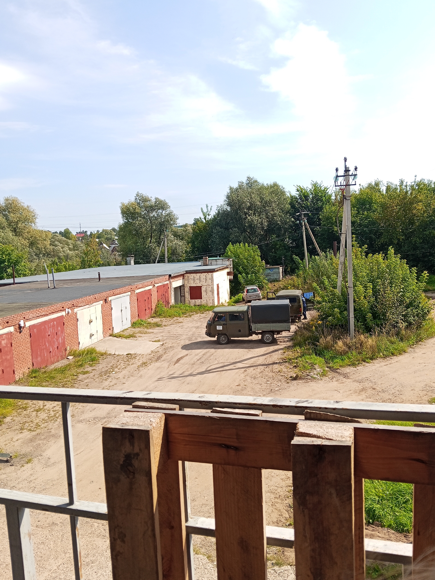The image depicts a view from an elevated position, looking out over a row of garages and a dirt road. In the foreground, a section of a wooden fence with metal railings is visible. Beyond the fence, a long, low building with a dark roof and brick facade stretches across the frame. This building has multiple garage doors, some red, some white, and one light gray, suggesting a storage or parking facility. Adjacent to the garages, a dirt road curves through the scene.
On the dirt road, a military-style truck with a canvas cover on its rear bed is parked. A second, smaller vehicle, possibly a car, is parked further down the road. A person is standing near the truck, and another is visible inside the cab of the truck. Trees and vegetation surround the area, with some taller trees and bushes forming a backdrop. Utility poles with electrical wires are also present in the middle ground, indicating the presence of infrastructure. The sky is partly cloudy with a bright blue hue.
The scene appears to be in a semi-industrial or residential fringe area, possibly a dacha community or a less developed part of a town. The presence of military-style vehicles and the utilitarian nature of the garages suggest a practical rather than a scenic location. The overall impression is one of an everyday, functional environment.
Given the details such as the architectural style of the garages, the type of vegetation, and the likely climate indicated by the clear sky and green foliage, it is plausible that this image was taken in Chaikovsky, Russia. Chaikovsky is known for its industrial areas, dacha settlements, and surrounding natural landscape, which are consistent with the elements observed in the photograph. The presence of utilitarian buildings like garages, along with the surrounding greenery and utility poles, aligns with typical scenes found in such Russian towns.