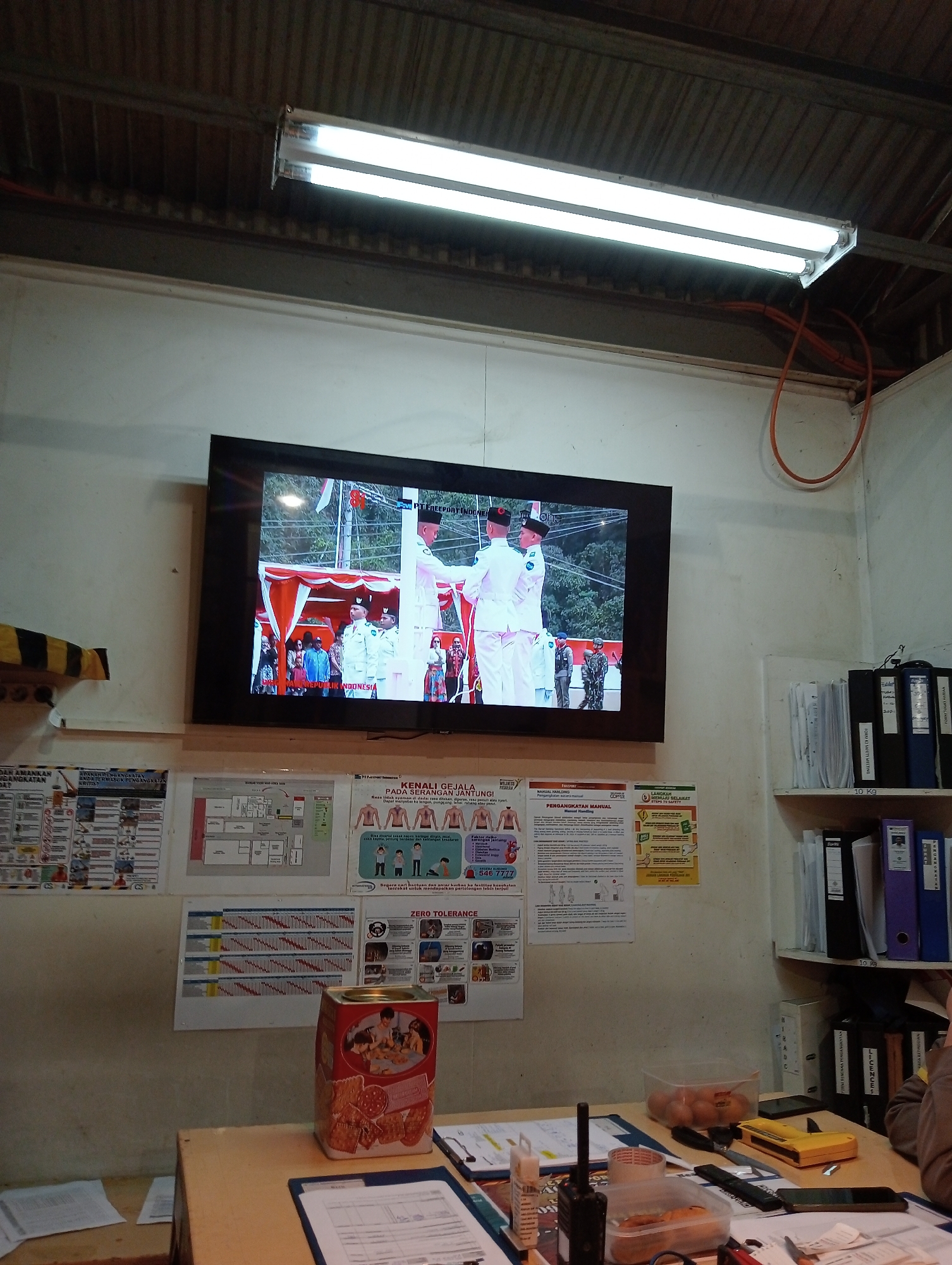 The image shows an indoor scene, likely an office or workspace, in Unknown, Indonesia. A large flat-screen television is mounted on the wall, displaying a broadcast of what appears to be a formal ceremony with people in white uniforms. Below the television, a bulletin board displays several posters with Indonesian text and diagrams, including one about heart attack symptoms and another detailing manual handling procedures. To the right of the bulletin board, a shelf holds several file binders and boxes. On the desk in the foreground, various items are visible, including a walkie-talkie, tape dispenser, food containers with fruit, a yellow electronic device, and stacks of papers. The environment appears functional and somewhat cluttered, with fluorescent lighting overhead and corrugated metal on the ceiling.