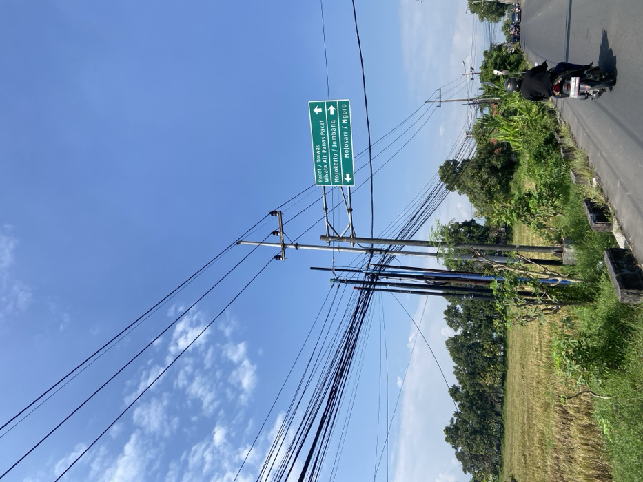 The image shows a rural road in Mojosari, Indonesia. The road is paved and has a single lane of traffic. A single motorcycle is traveling towards the camera. To the right of the road is a field of tall grass. To the left of the road is a grassy area with trees. The road is lined with several power poles and utility lines. One power pole has a sign with the following text: "Pacet / Trawas, Wisata Air Panas Pacet, Mojokerto / Jombang, Mojosari / Ngoror". The sky is blue with a few clouds. The weather is clear and sunny. The coordinates of the image are -7.503 latitude and 112.531 longitude.