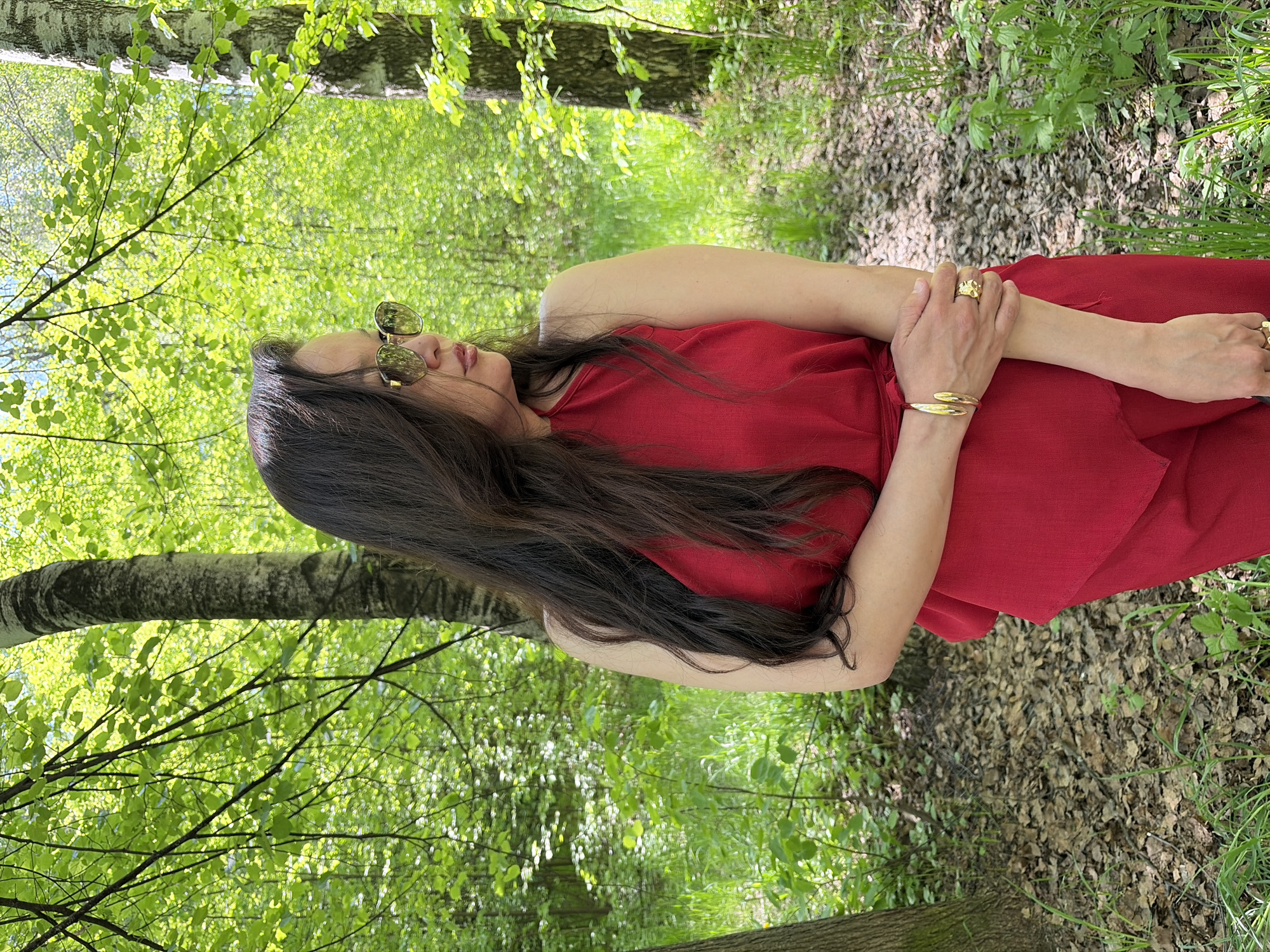 A woman in a red dress stands in a forest of birch trees. She wears sunglasses and gold jewelry. The forest floor is covered with leaf litter and green plants.
