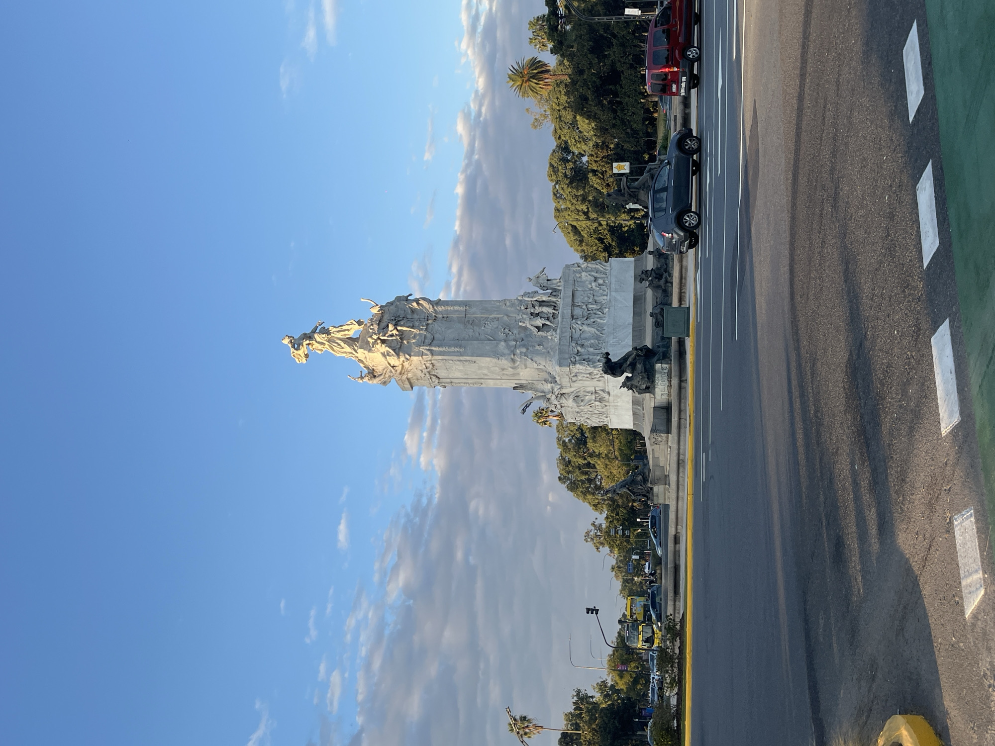 An upright, sunlit photograph captures a grand stone monument in a city environment, likely in Buenos Aires, Argentina. The monument, positioned on the left side of the frame, is elaborately sculpted with numerous classical figures and relief carvings, appearing in a light, possibly white or cream-colored stone. A prominent group of statues crowns its uppermost section.
To the right of the monument, a wide asphalt road stretches into the distance, marked with bright yellow lines and white rectangular pavement markings, including a distinctive green-painted section on the far right, indicative of a bike lane. Several vehicles are visible: a dark-colored hatchback car is parked alongside the monument, a red van is seen further back amongst the trees, and multiple yellow double-decker buses (likely tourist vehicles) are visible down the road. Traffic lights can also be seen in the distance.
The background features a clear blue sky with scattered white clouds on the left. On the right, behind the road and cars, a dense cluster of lush green trees, including some palm trees, creates a natural backdrop.
The lighting suggests either late afternoon or early morning, with warm, golden sunlight illuminating the monument and casting long shadows across the road, indicating a low sun angle. The scene is quiet, with no specific activity beyond the typical presence of vehicles and a standing monument. No discernible text is visible on signs or buildings.