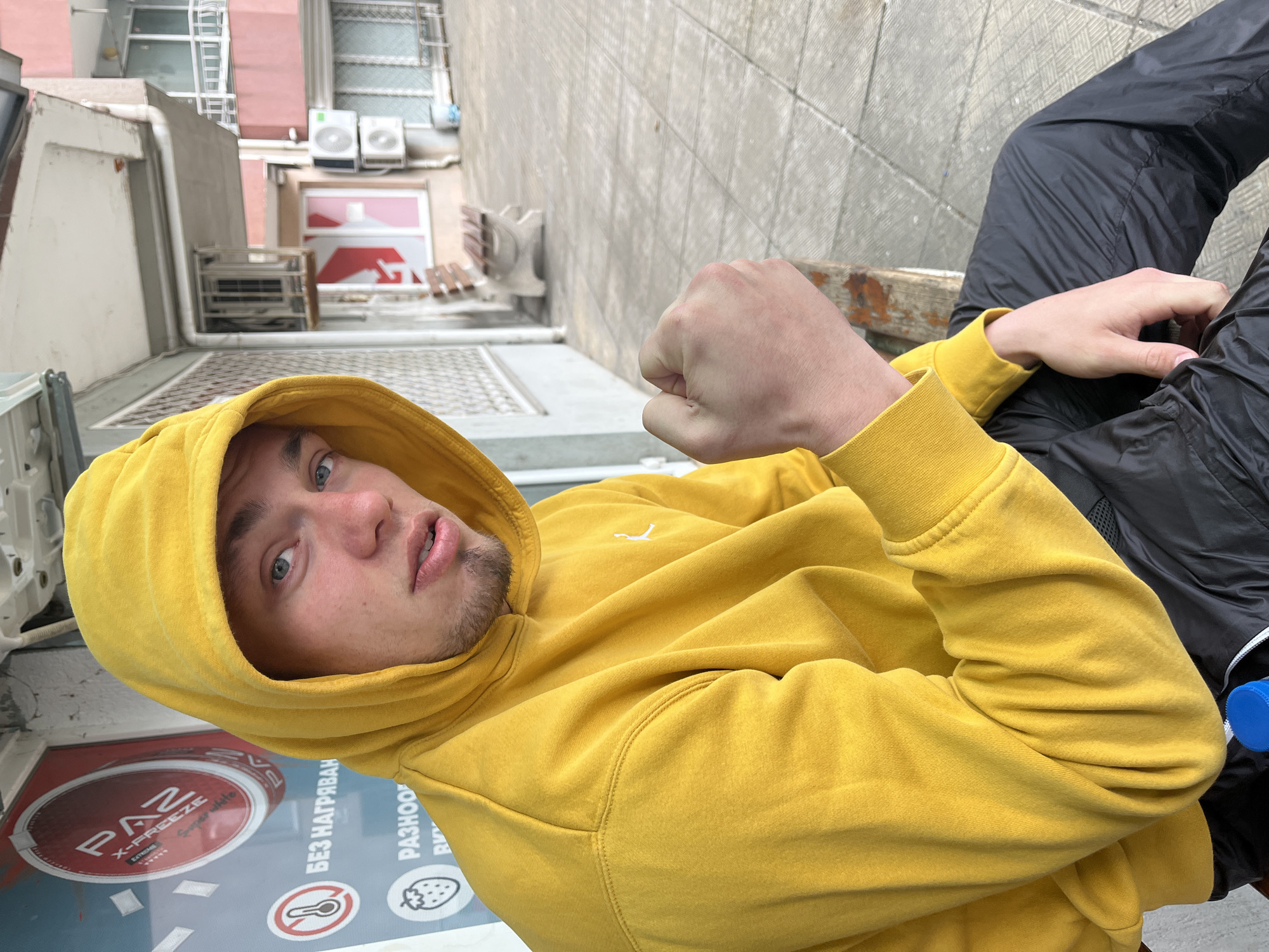 A medium, eye-level shot of a young man in a yellow hooded sweatshirt sitting on a bench, likely in Burgas, Bulgaria, judging by the Cyrillic writing. He wears dark-colored track pants and has a close fist raised to chest level. The background shows buildings and alleyways, typical of an urban setting. In the lower-left is an advertisement sticker. The man looks at the camera.