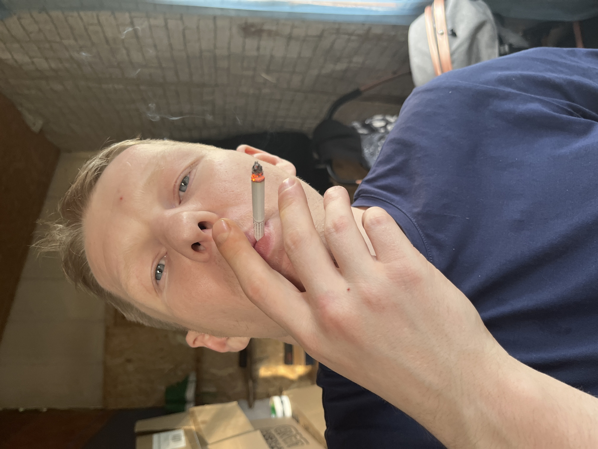 A fair-skinned person with short, light blonde hair is holding a lit cigarette to their lips. The person is wearing a dark blue shirt. Smoke is visible rising from the cigarette. The background shows what appears to be a room with brick or block walls and various objects, including a grey bag with orange straps, boxes, and other indistinct items. No identifiable location in the city of Vesyoloye, Russia, is discernible from the image.
