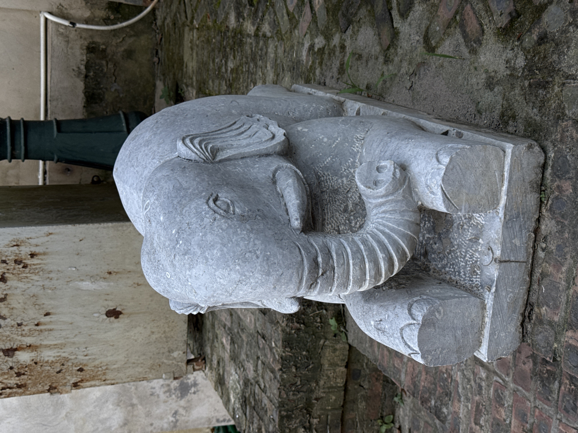A stone sculpture of an elephant rests on a base. The sculpture is light gray and appears aged. The elephant is depicted lying down, with its trunk curled upwards and its legs folded. It is placed on a weathered stone base, which sits on what looks like a brick or stone surface, likely a pathway or a low wall. The background includes greenery and what appears to be a building wall or fence. The location is specified to be Hà Nội, Vietnam.