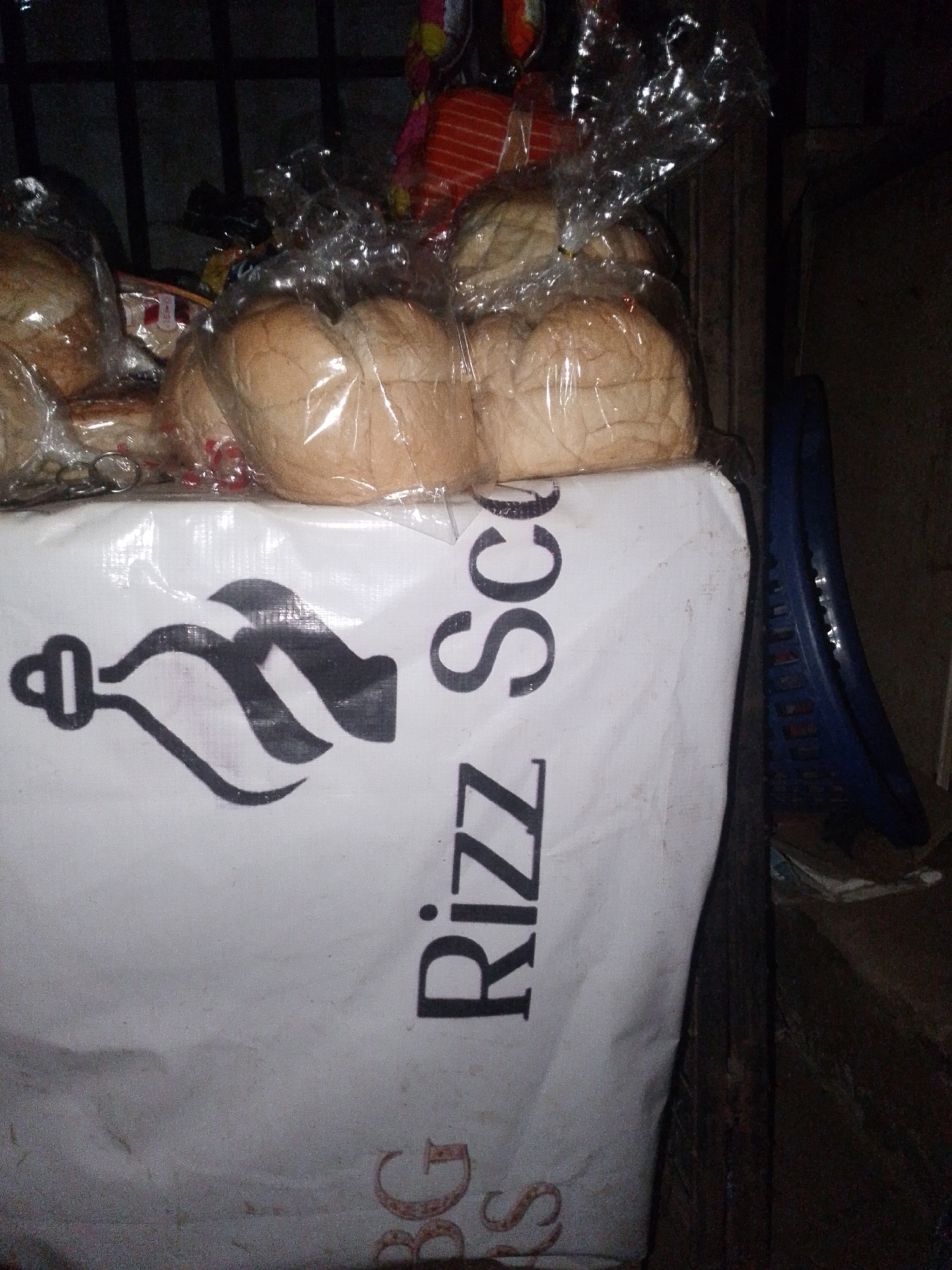 The image shows a stack of bread loaves in clear plastic packaging on top of a white bag with the words "Rizz Sco". The bag also has a black logo. The location appears to be a small store or market in Ifite-Awka, Nigeria, judging by the type of goods and the general environment, which includes a blue plastic object adjacent to the bag. The lighting is dim.