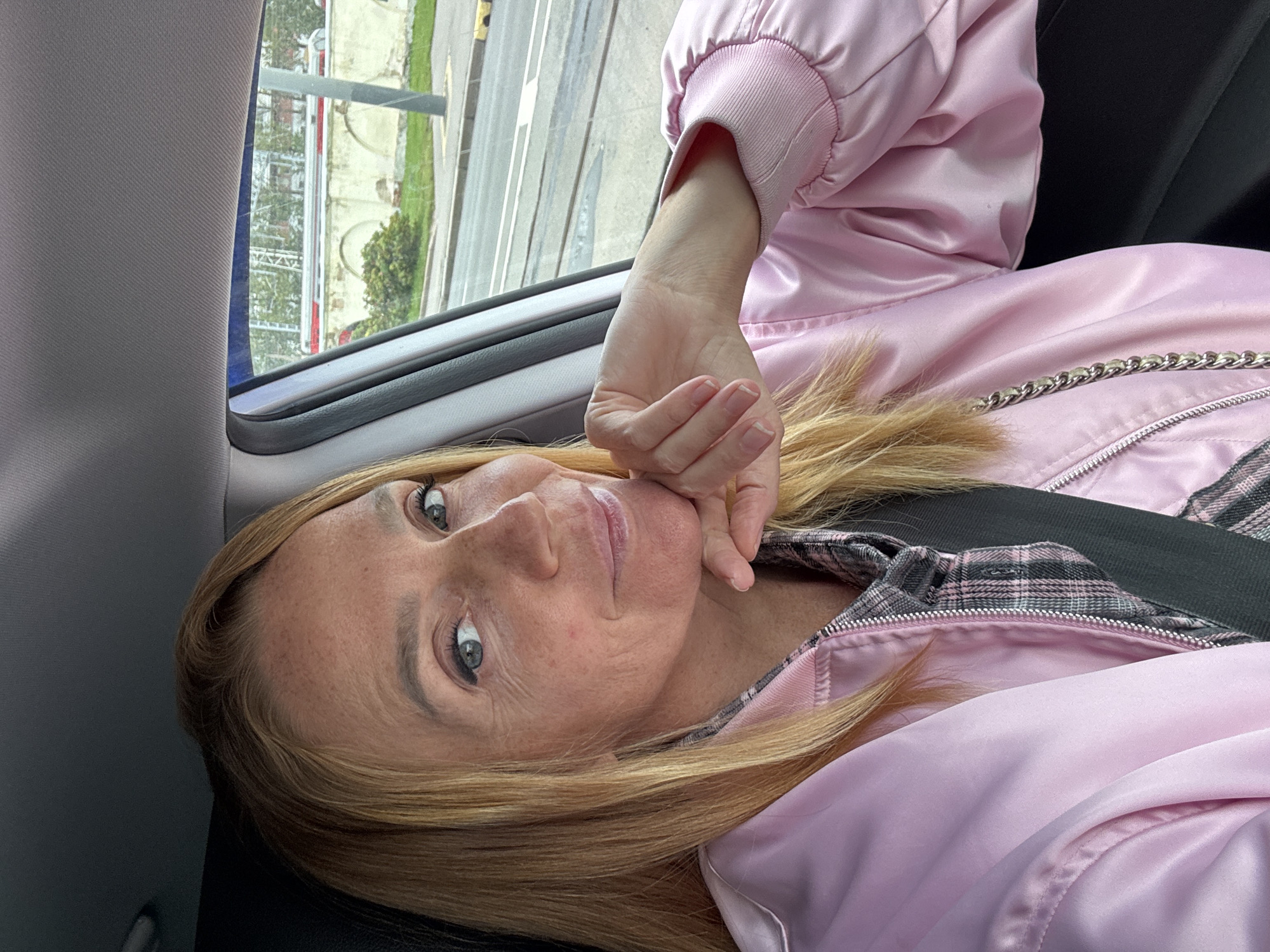 A blonde woman is seated in the driver's side of a car, taking a selfie. She is wearing a pink satin bomber jacket over a grey and pink plaid shirt. Her right hand is resting on her chin, and she is looking at the camera with a slight smile. Through the car window, a street scene in Saint Petersburg, Russia, is visible, featuring tram tracks, a sidewalk, and buildings with arched windows. The lighting suggests it is daytime.