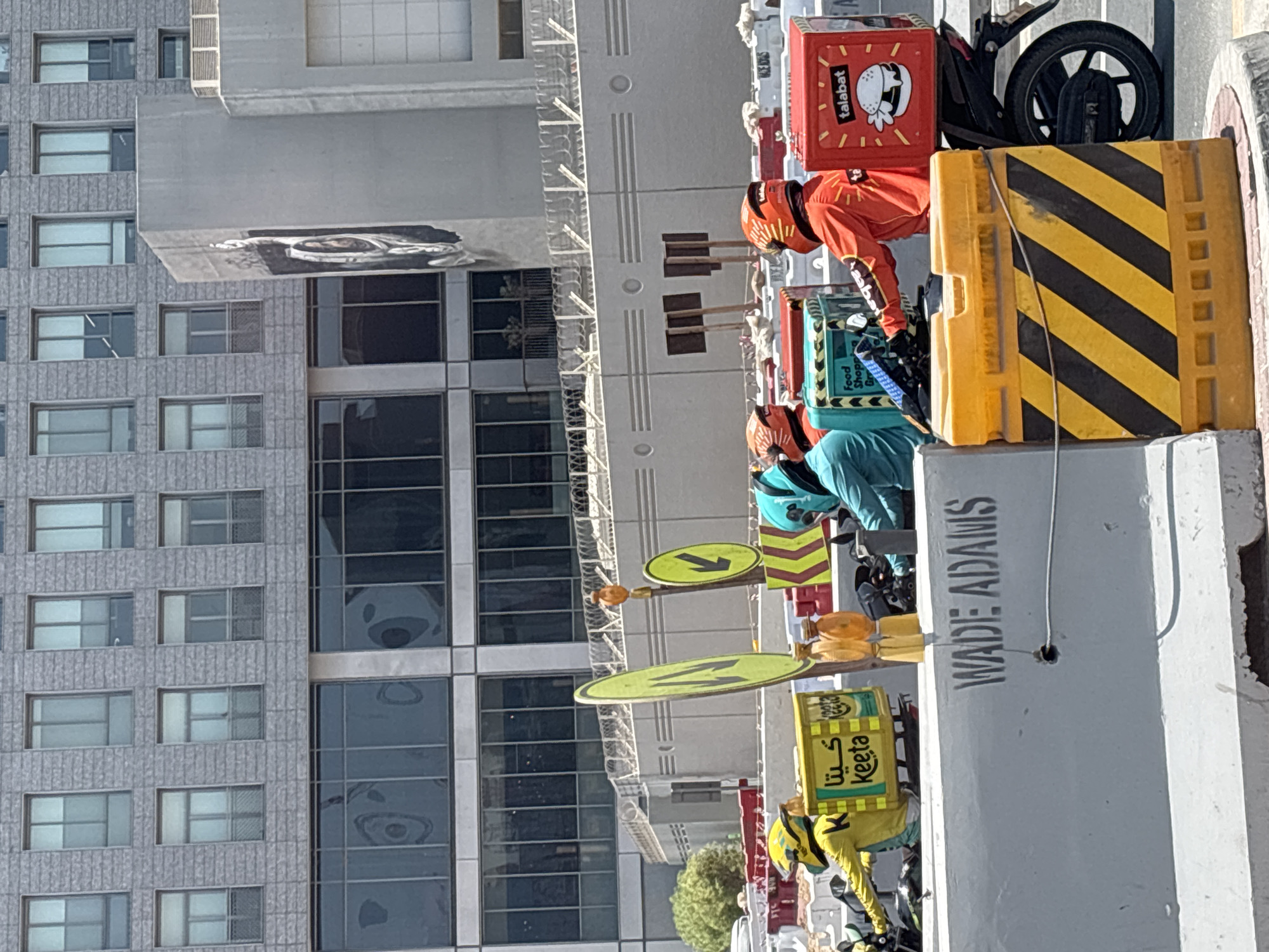 The image depicts an outdoor scene in Dubai, United Arab Emirates, likely a street or construction area. In the foreground, several delivery riders on motorcycles are present. One rider, wearing a yellow jacket and helmet, has a yellow delivery box labeled "Keeta." Another rider in an orange jacket and helmet is positioned next to a red delivery box labeled "talabat." A third rider in a blue outfit and helmet is also visible. The riders appear to be waiting or preparing for deliveries.
Behind the riders, there are several construction barriers, including yellow and black striped traffic barriers and concrete barriers. One concrete barrier is stenciled with "WADE ADAMS." In the mid-ground, there are signs, including a yellow directional sign with an arrow. A low-rise building with barred windows and decorative elements is situated behind the barriers and signs.
The background is dominated by a modern, multi-story building with a facade of gray stone and numerous windows. The upper portion of the building has regularly spaced rectangular windows, some of which appear to be frosted or tinted. The lower section of this building features large, dark-tinted windows. A piece of public art or mural, depicting a face or figure, is visible on the side of a grey structure to the right of the main building. The overall impression is a blend of urban infrastructure, commercial activity, and ongoing development.