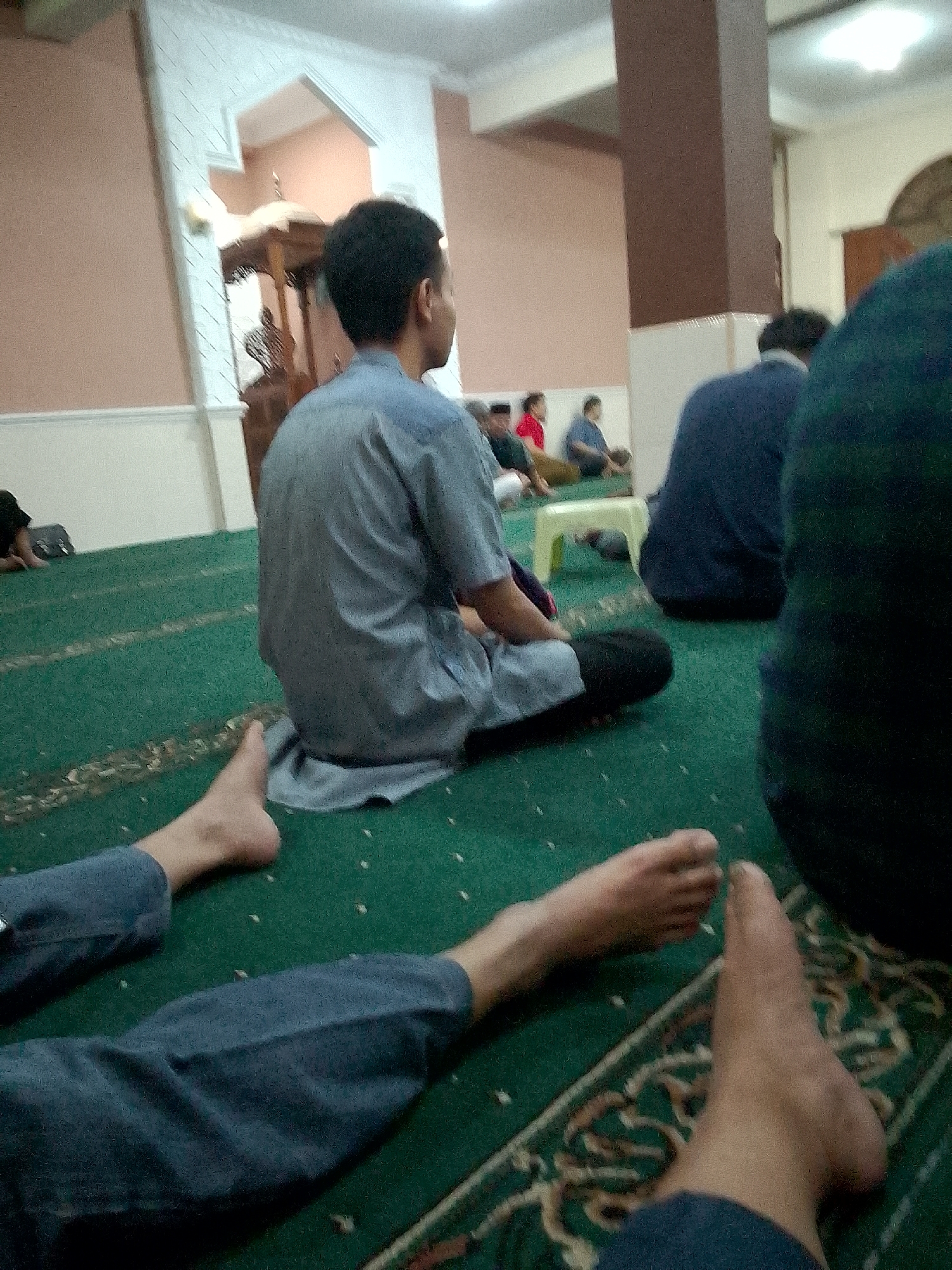 The image shows the interior of a mosque in Kertajaya, Indonesia, with several people present. In the foreground, two pairs of bare feet are visible, suggesting people are sitting on the floor. The floor is covered with a green carpet with a patterned design. In the mid-ground, a man in a light blue, collared shirt is seated on the carpet, with his back to the camera. He appears to be wearing dark pants. Behind him, other individuals are seated on the floor, including a person in a dark blue top and another in a dark green top. Further in the background, a wooden pulpit is visible, a common feature in mosques. The mosque's architecture includes arched doorways and textured walls in shades of pink and white. The lighting suggests an indoor environment, possibly during prayer time.