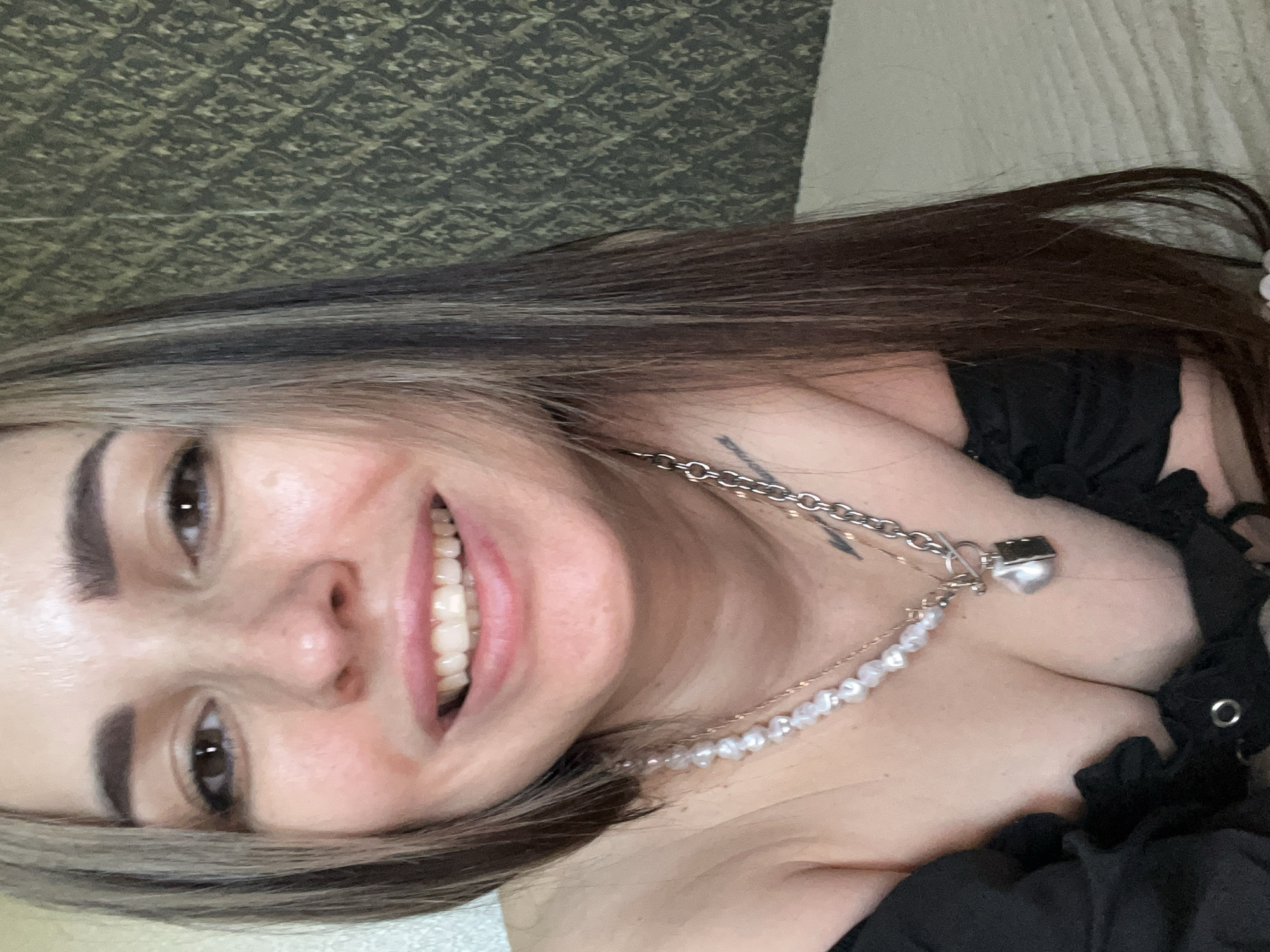 A young woman smiles at the camera. She has long dark hair with blonde streaks and light skin. She wears a black top and two necklaces: one silver chain with a padlock pendant and one strand of pearls. A tattoo is visible on her upper chest. The background consists of a patterned wall and a white surface, possibly a bed. The location and city are unknown.