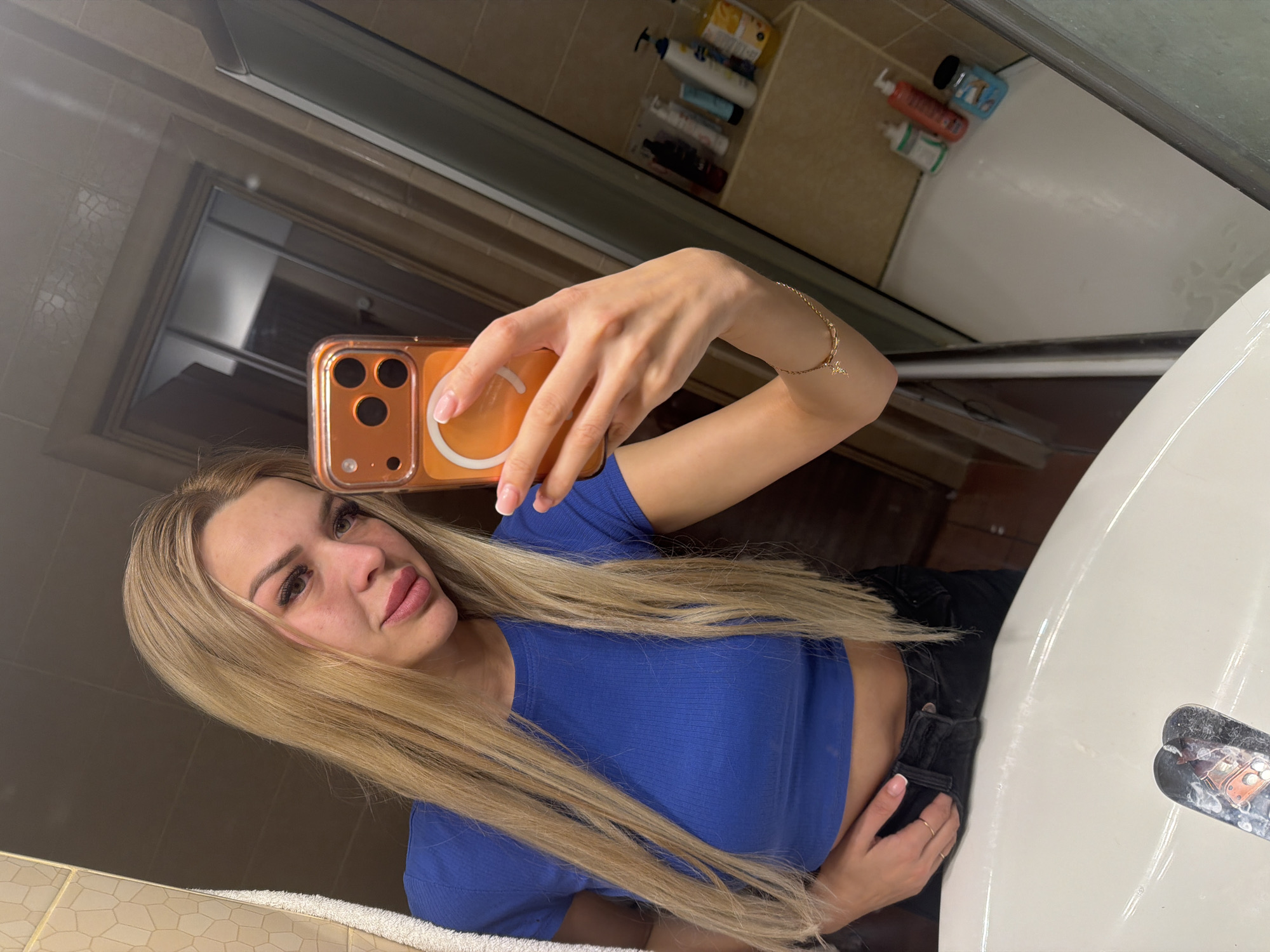 A blonde woman with long, straight hair takes a mirror selfie in a bathroom. She is wearing a blue crop top and black pants. Her phone, with an orange case, is visible in her hand. The bathroom appears to have tiled walls and a shower stall with various bottles of toiletries. The sink is in the foreground of the image.