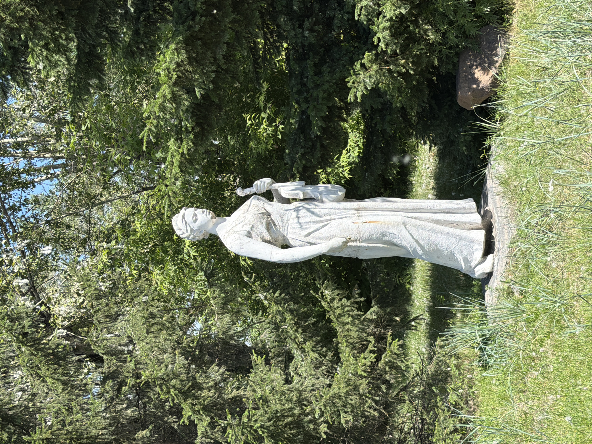 The image shows a full-body white stone statue of a woman standing in a park. The statue is located in the city of Krasnoyarsk, Russia, specifically in the Central Park of Culture and Recreation. The woman is depicted wearing a long, flowing dress and holding a staff in her right hand, with a draped cloth or garment in her left arm. She is positioned in front of a dense backdrop of evergreen trees and other foliage, with sunlight filtering through the leaves. The ground in front of the statue is covered with green grass, with some areas appearing more overgrown and wild. A large, irregularly shaped rock is visible to the right of the statue, partially shaded by the surrounding greenery. The overall scene suggests an outdoor public space, likely a garden or park area, with the statue serving as a decorative or memorial element.