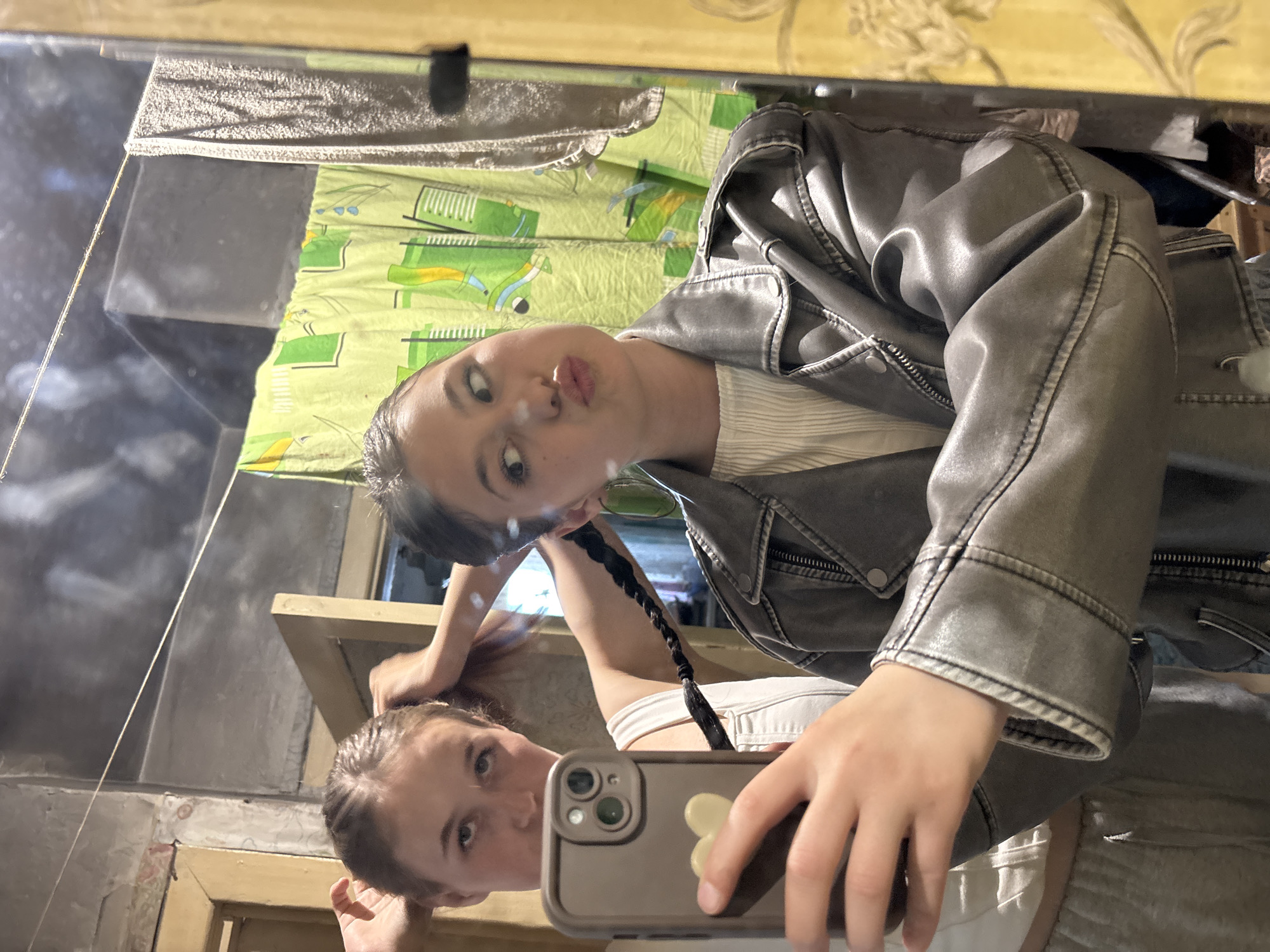 A selfie is taken in a mirror showing two reflections of the same young woman. In the primary reflection, she is wearing a grey jacket, a striped shirt, and has a braid. She is posing with pursed lips. The second reflection shows her holding the phone, wearing a white top and adjusting her hair. The background includes a green and yellow patterned cloth and parts of a room with painted walls. There is no verifiable evidence to suggest the location is in the city of Фоки, Russia.