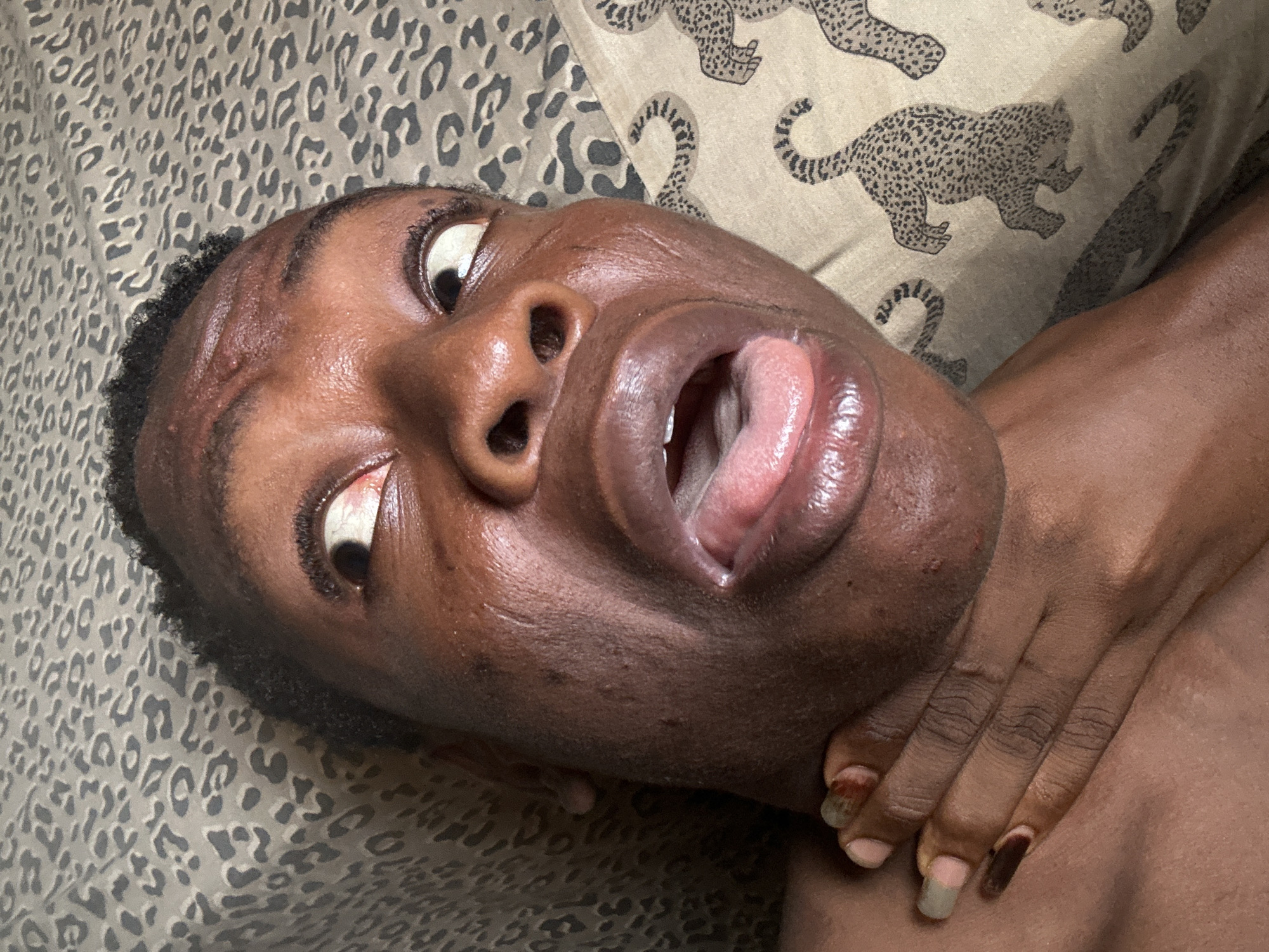A close-up, angled shot features the face and upper body of a person with dark skin and short, dark, curly hair. The person's head is tilted to the side, and their eyes are wide open, rolled upwards to prominently display the white sclera. Their mouth is open, with the tongue extended slightly, revealing the pink interior.
A hand with dark skin is positioned around the person's neck, with fingers gently grasping or resting on the left side of the neck. The fingernails of this hand are painted, some a light, muted color (possibly pale gray or green), and others a darker shade (possibly dark brown or maroon), with some visible growth at the cuticle area.
The person's skin shows some texture, with small bumps visible on the forehead and cheeks. The background consists of a patterned fabric, likely a bedsheet or pillowcase, featuring a stylized leopard or cheetah print with various silhouettes of big cats in motion. The fabric's colors are muted, primarily beige/tan with dark gray/black patterns. Part of the person's bare shoulder or chest is visible beneath the hand. The lighting is soft and even, highlighting the features without harsh shadows. There is no visible text in the image.